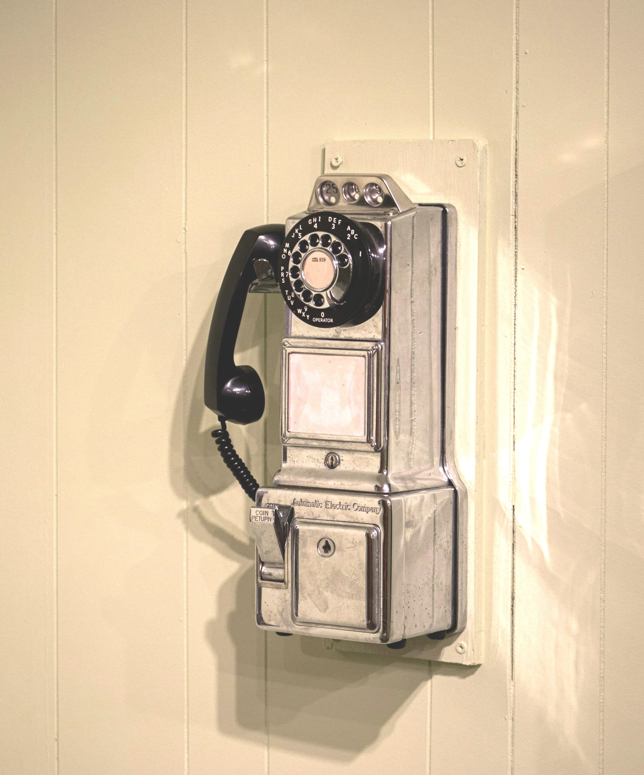 A vintage payphone mounted on a plain beige wall, featuring a rotary dial, coin slots, and a metal casing.