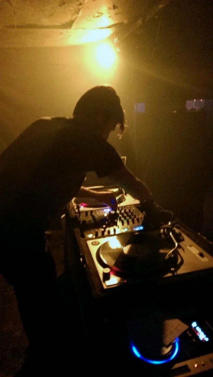A DJ with wearing a black shirt playing music on a turntable in a dimly lit club with yellow lighting.