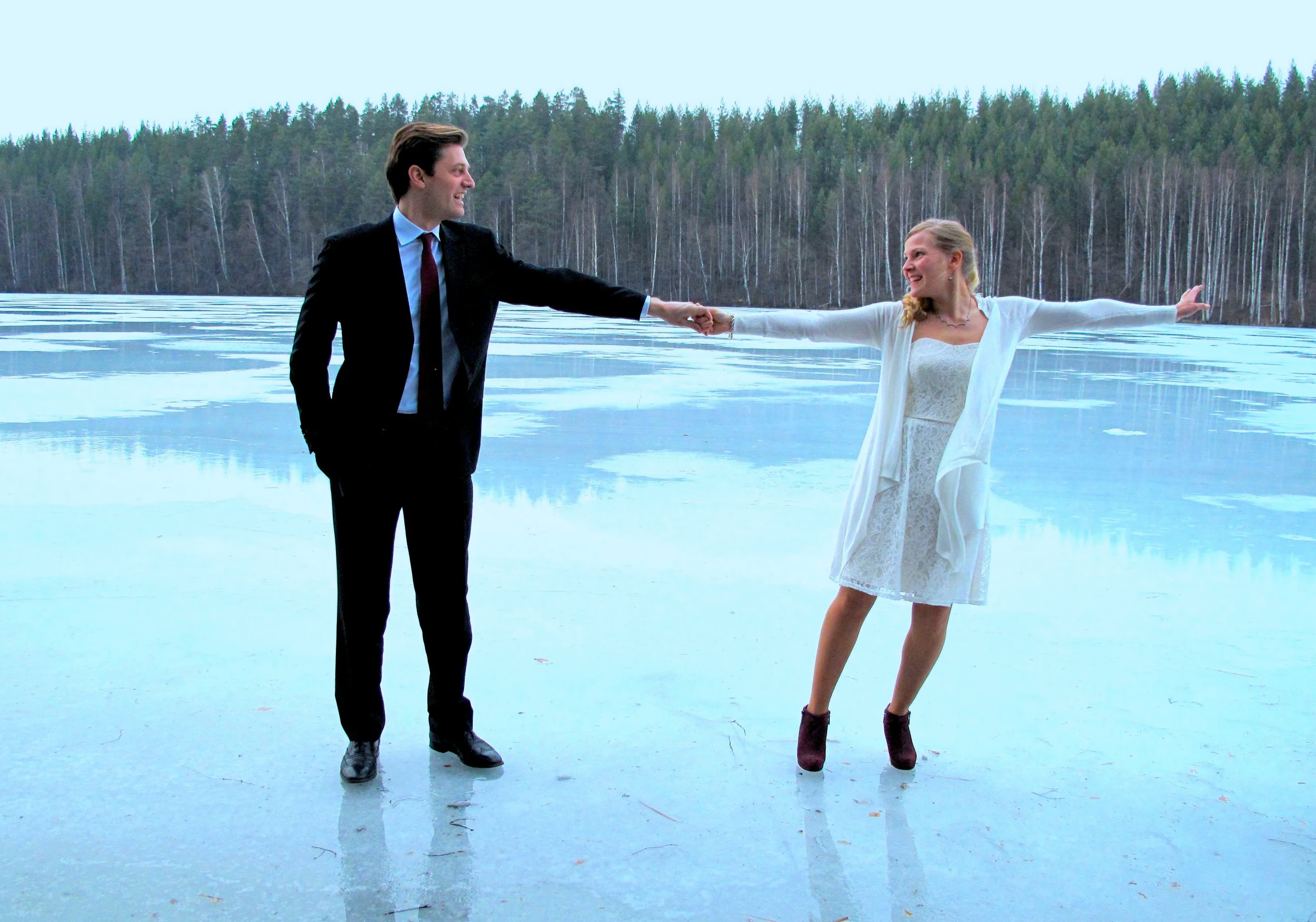A young man and woman are standing on a frozen lake surrounded by trees. The man is wearing a suit and tie while the woman is in a lace dress with a white coat. They are holding hands and smiling at each other having just married.