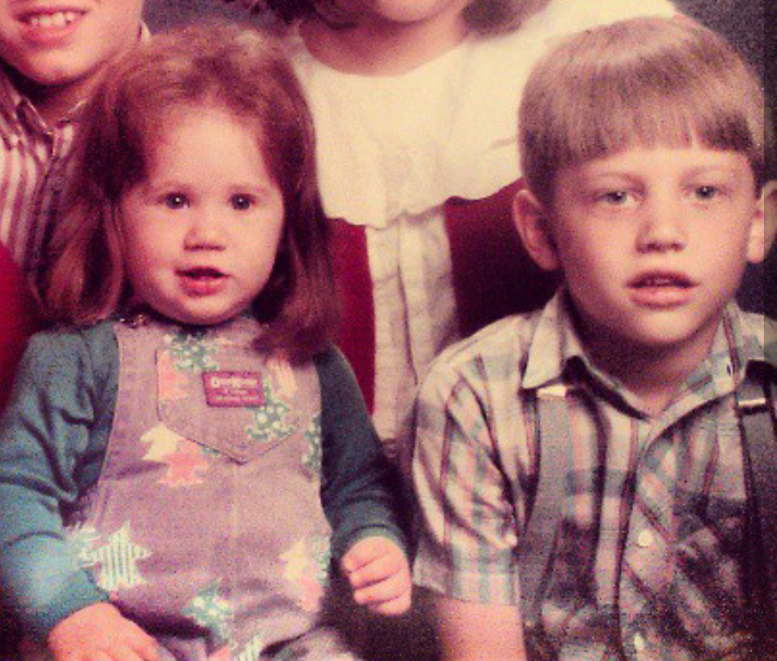 A young girl with red hair and a boy with blonde hair sitting together, part of a group photo with other children partially visible in the background.