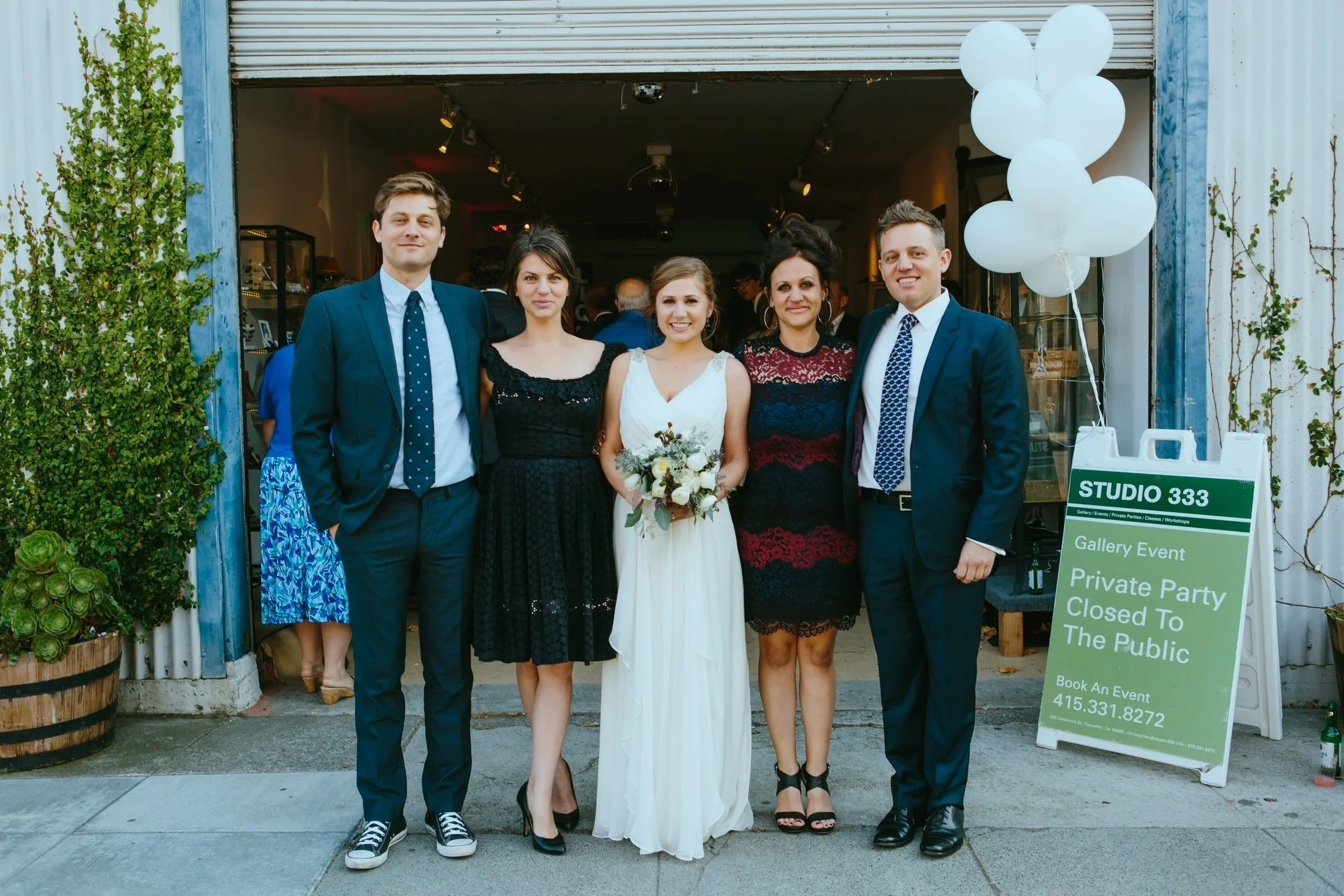 Group of five people standing outside a gallery or event space, including a bride holding a bouquet, two women, and two men in suits, with balloons and signage for a private event.