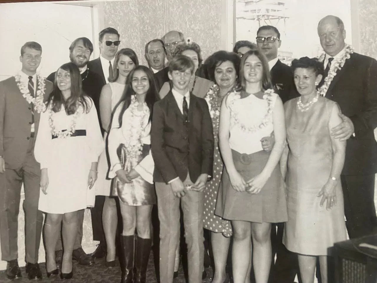 A group of people celebrating at a gathering, mostly women wearing leis, with a few men dressed in suits, some wearing glasses, and one man in a tuxedo, posing for a photo indoors.