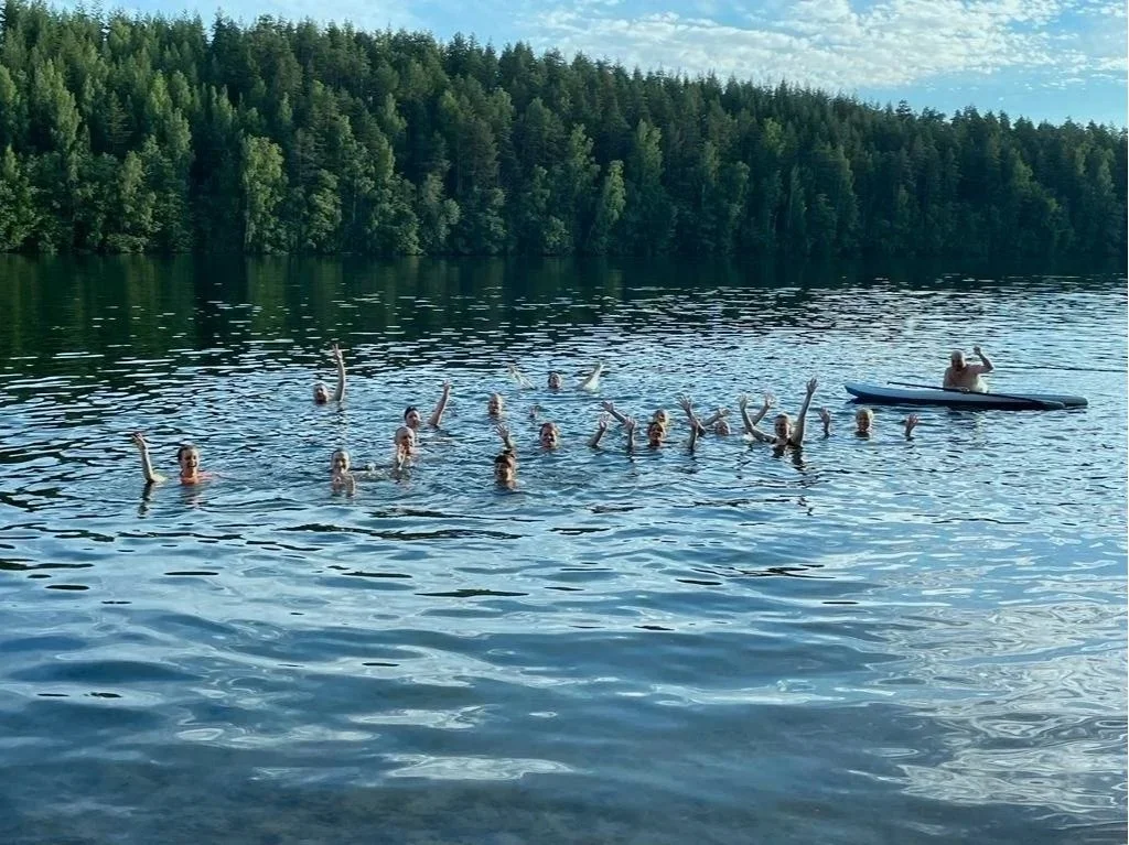 A group of people swimming at a lake party with lush green trees and a partly cloudy sky in the background, and a person on a kayak to the right.