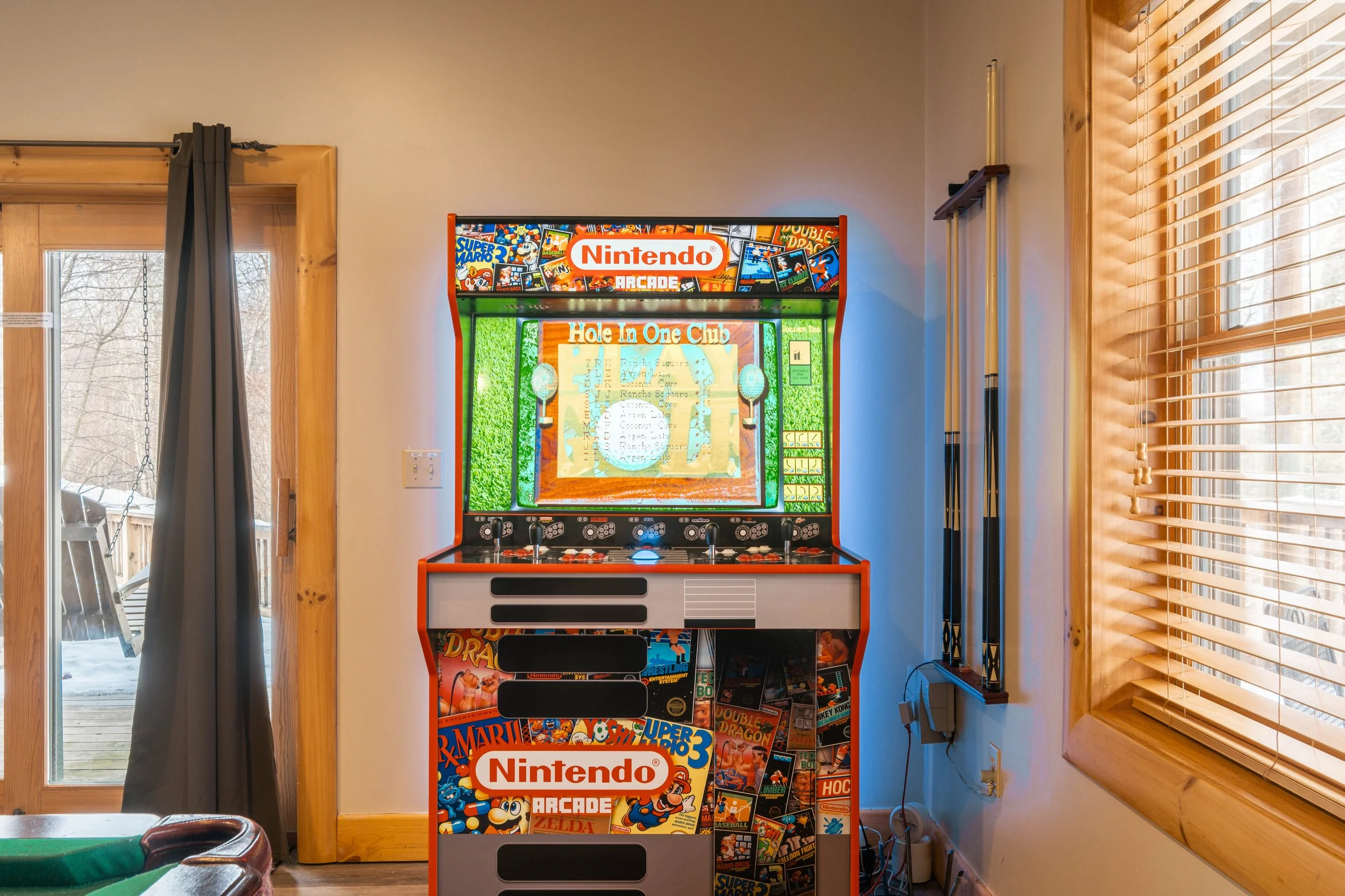 A vintage Nintendo arcade game machine with colorful graphics and posters, situated indoors next to a window with blinds. Outside, there is snow on the deck, and on the left, a sliding glass door has a curtain.