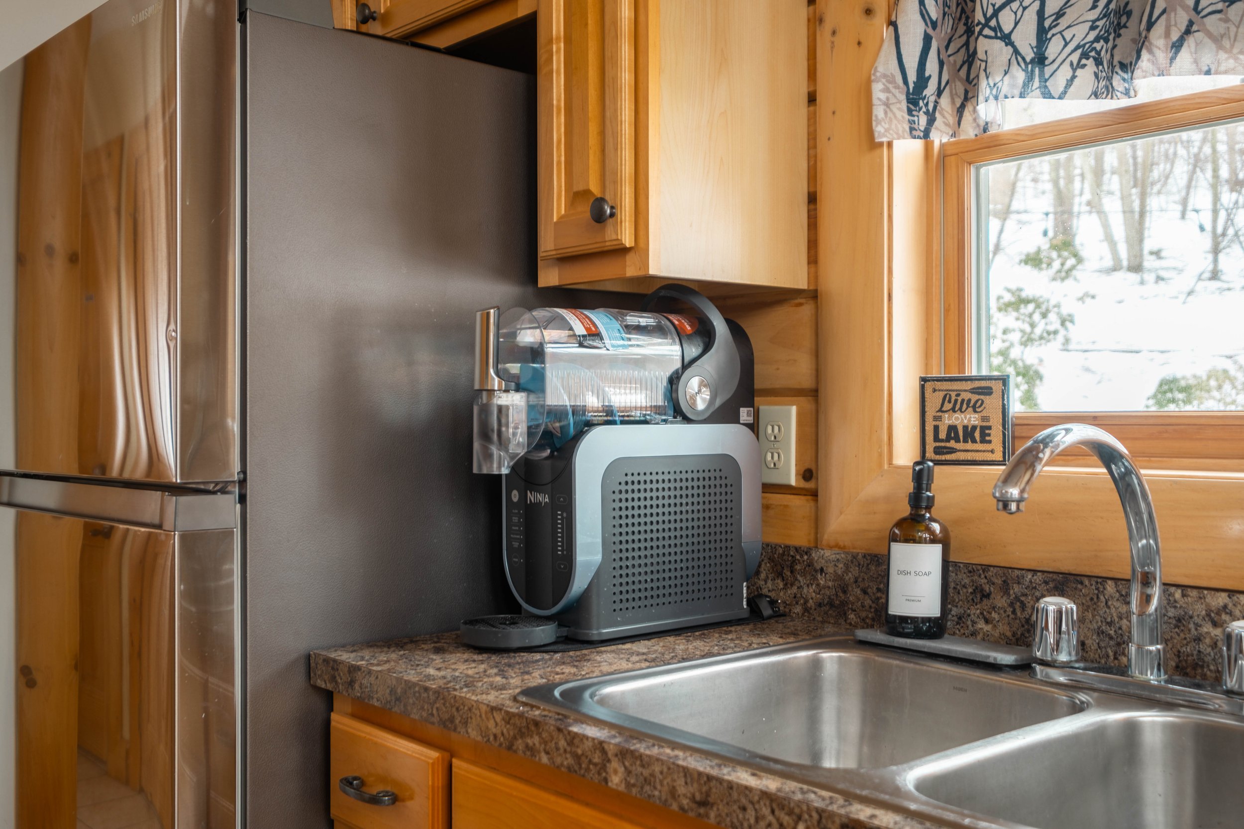 Kitchen countertop with a coffee maker, water filter, dish soap, and a window showing snowy trees outside.