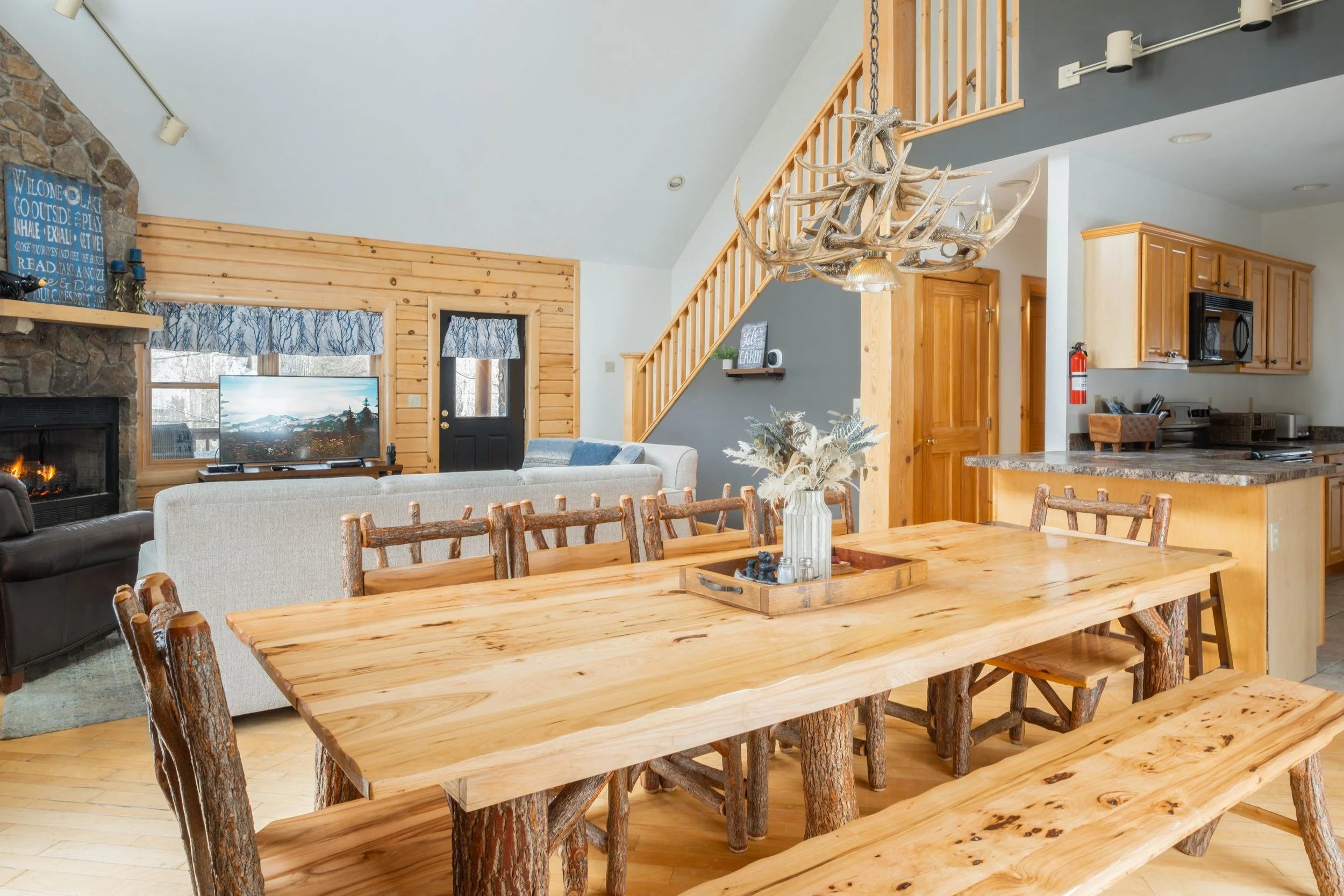 Open-concept living and dining room with wooden accents, a large dining table made of rustic wood, a staircase with wooden railing, a kitchen with light wood cabinets, and a stone fireplace.