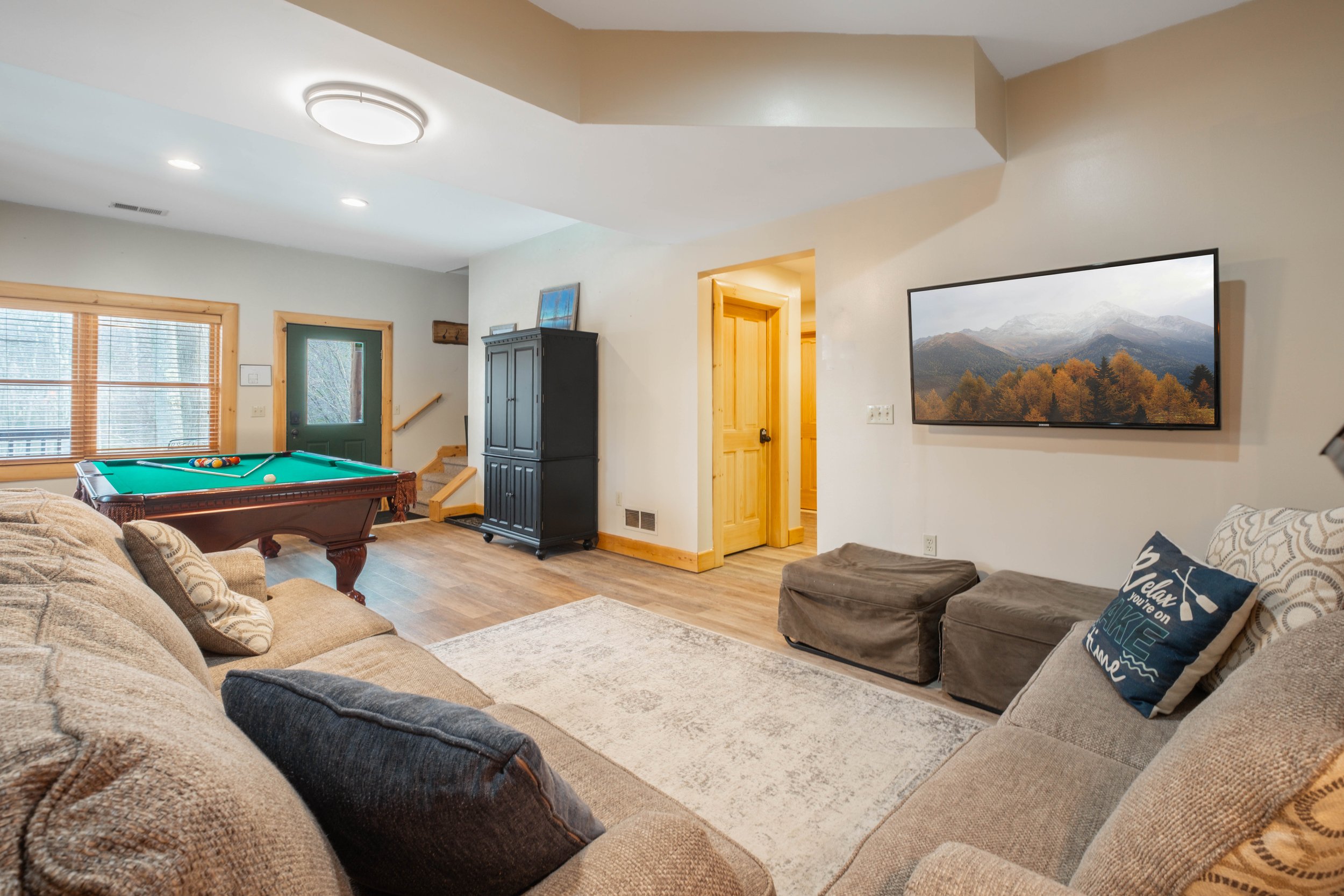 Living room with a beige sectional sofa, a pool table, a television on the wall, and a wooden door leading to another room.