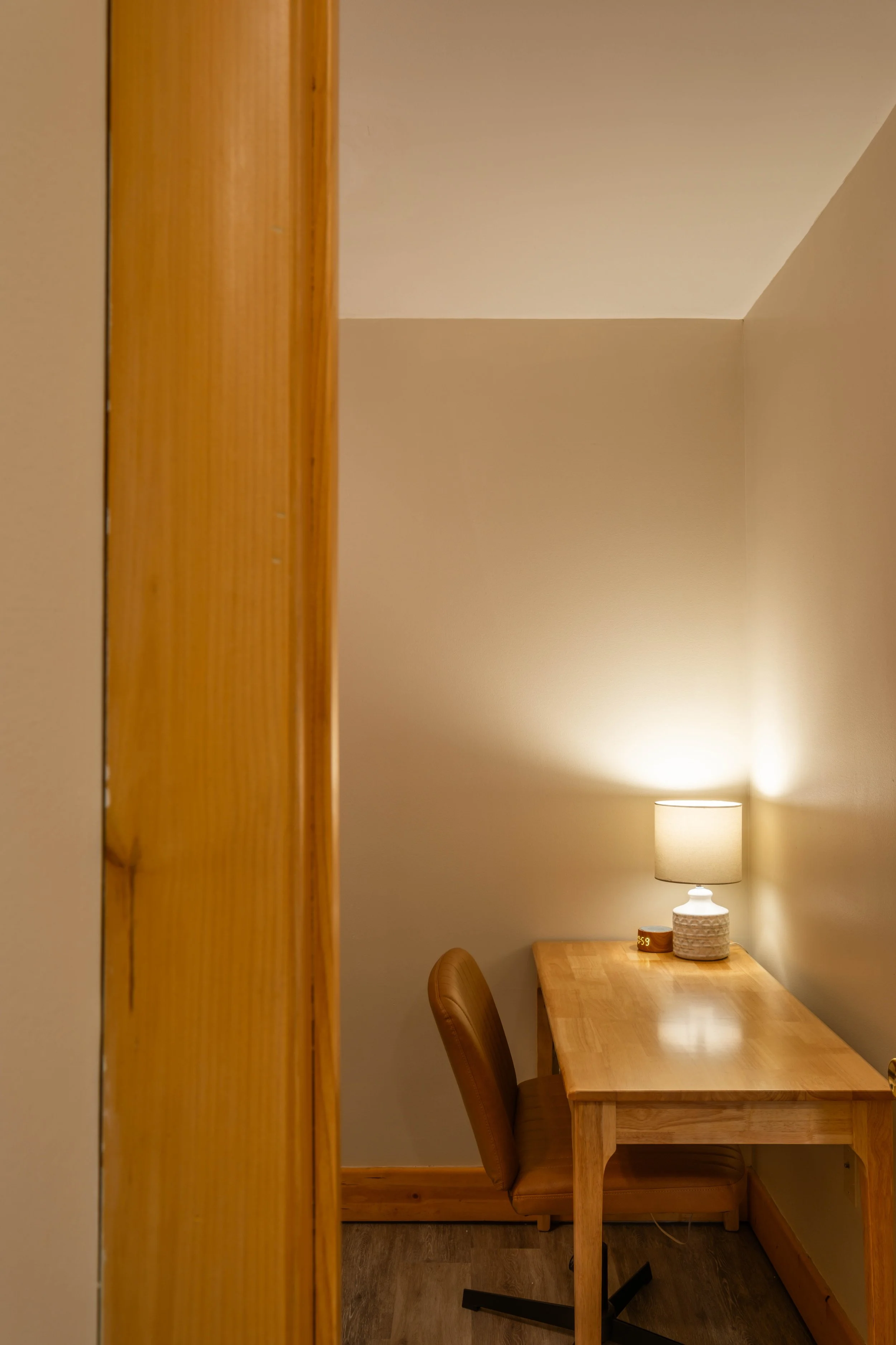 A small wooden desk with a lamp and an alarm clock, next to a brown leather office chair.