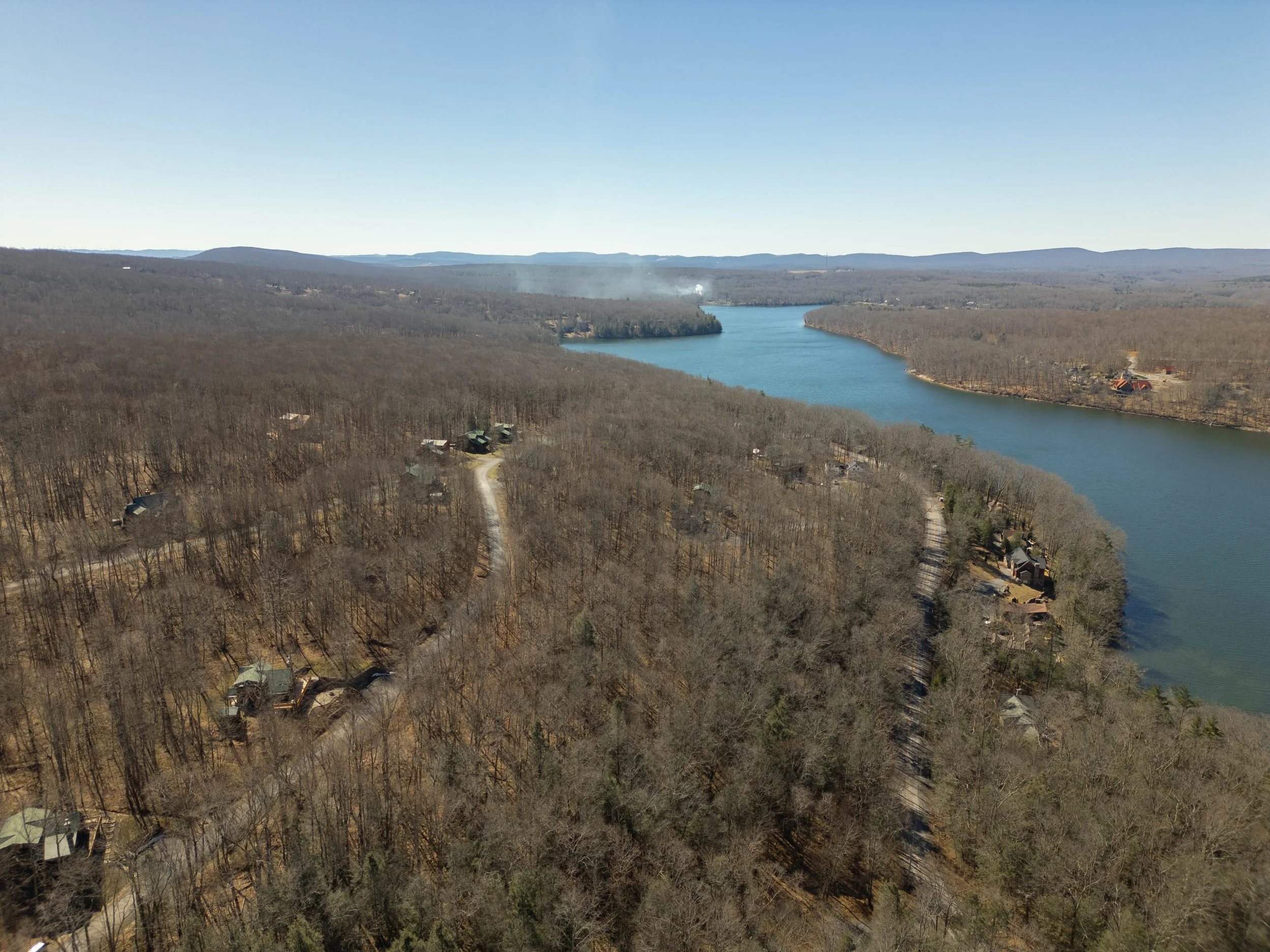 Aerial view of a river flowing through a forested landscape with scattered houses and a distant mountain range under a clear sky.