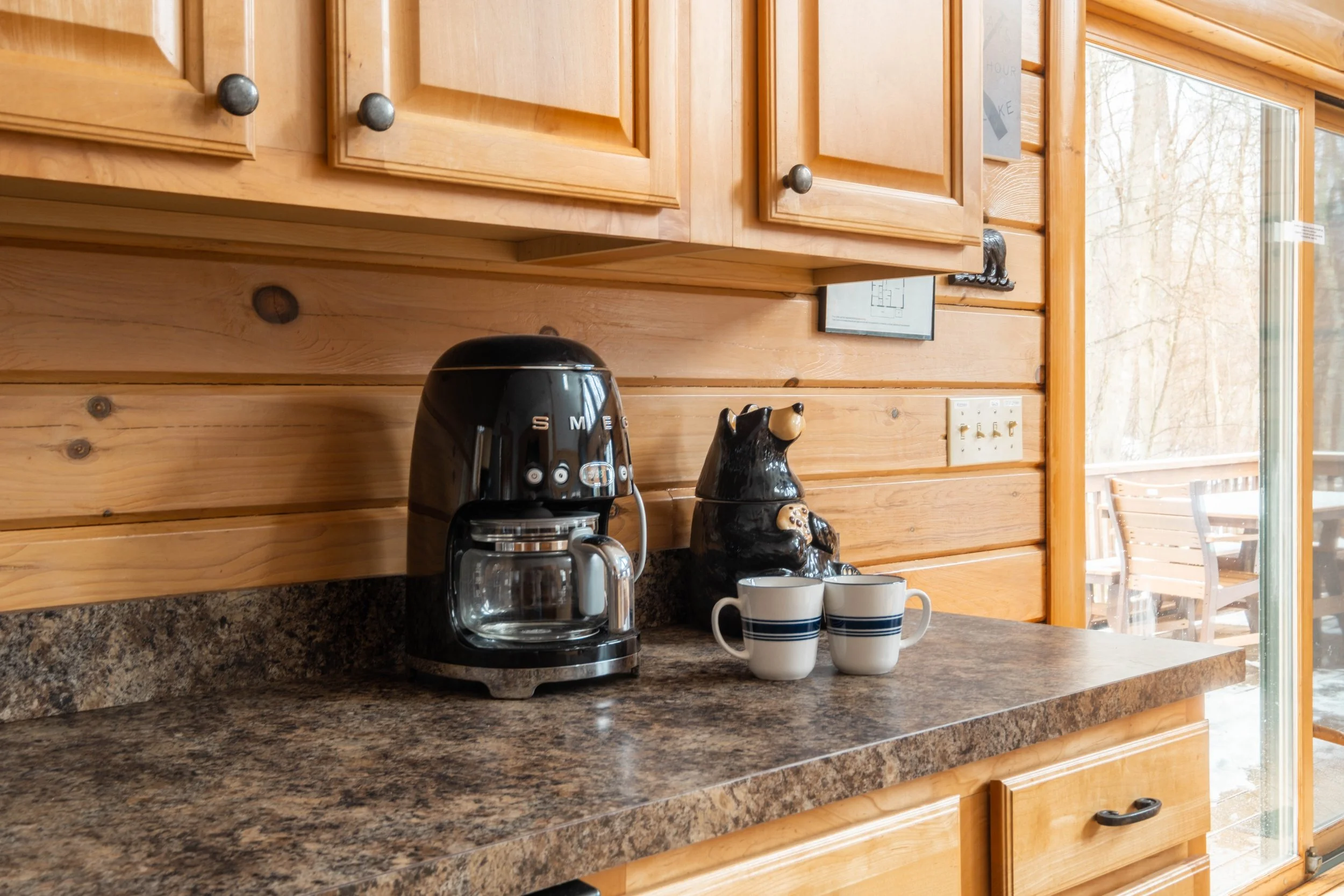 Coffee maker, bear-shaped ceramic figurine, and two striped coffee mugs on a granite kitchen countertop in a wooden kitchen with a large glass door leading to an outdoor seating area.
