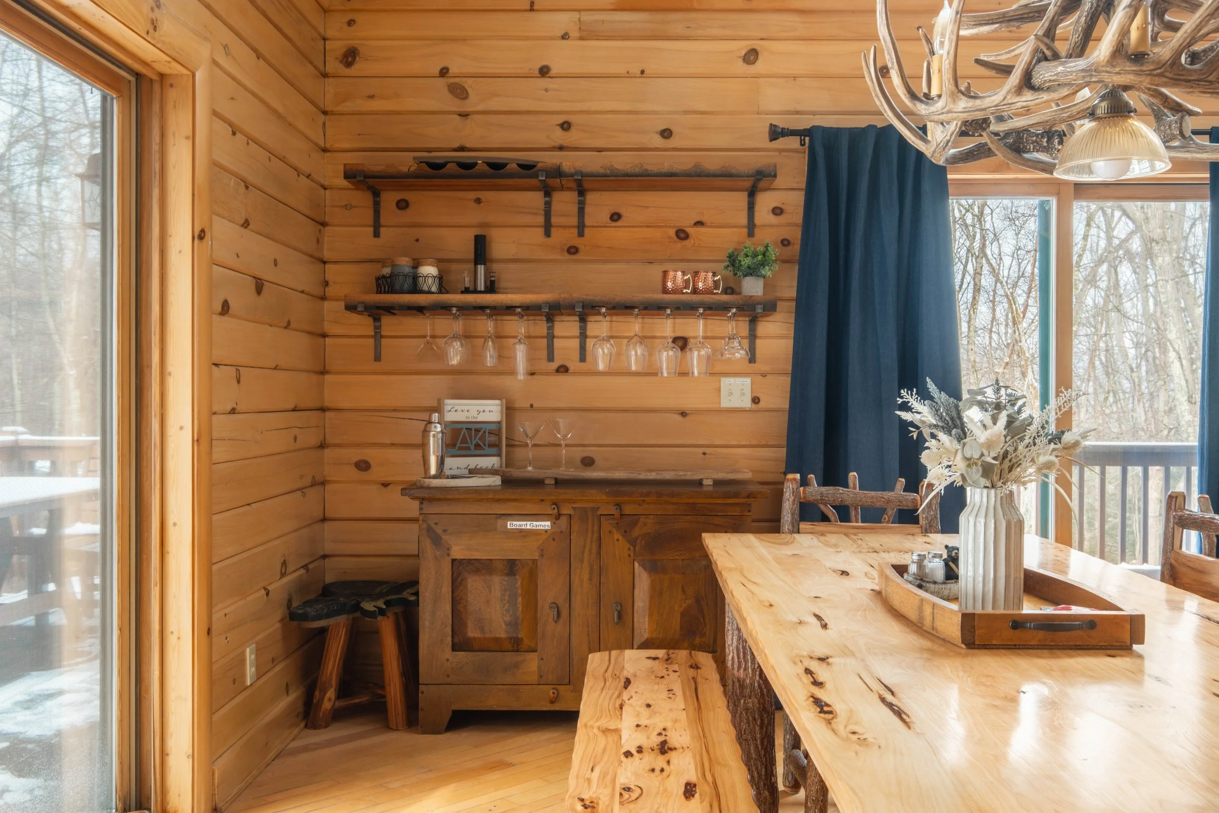 Wood-paneled dining area with a large wooden table, a vase with flowers, and a sideboard. There is a window with blue curtains revealing an outdoor winter scene. A rustic chandelier made of antlers hangs above the table.