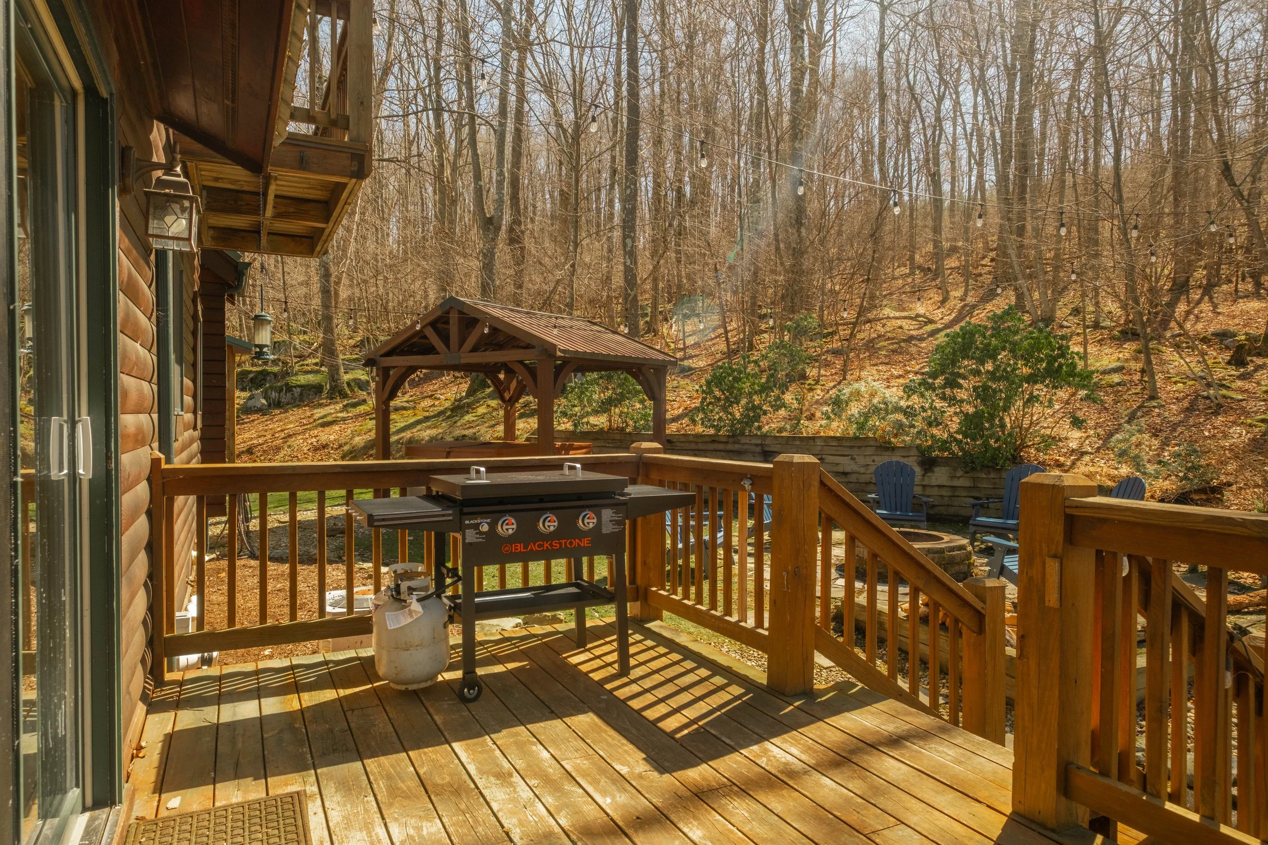 Wooden deck attached to a house, featuring a gas grill on wheels, a fire pit with blue chairs around it, and surrounded by leafless trees and a wooded hillside.