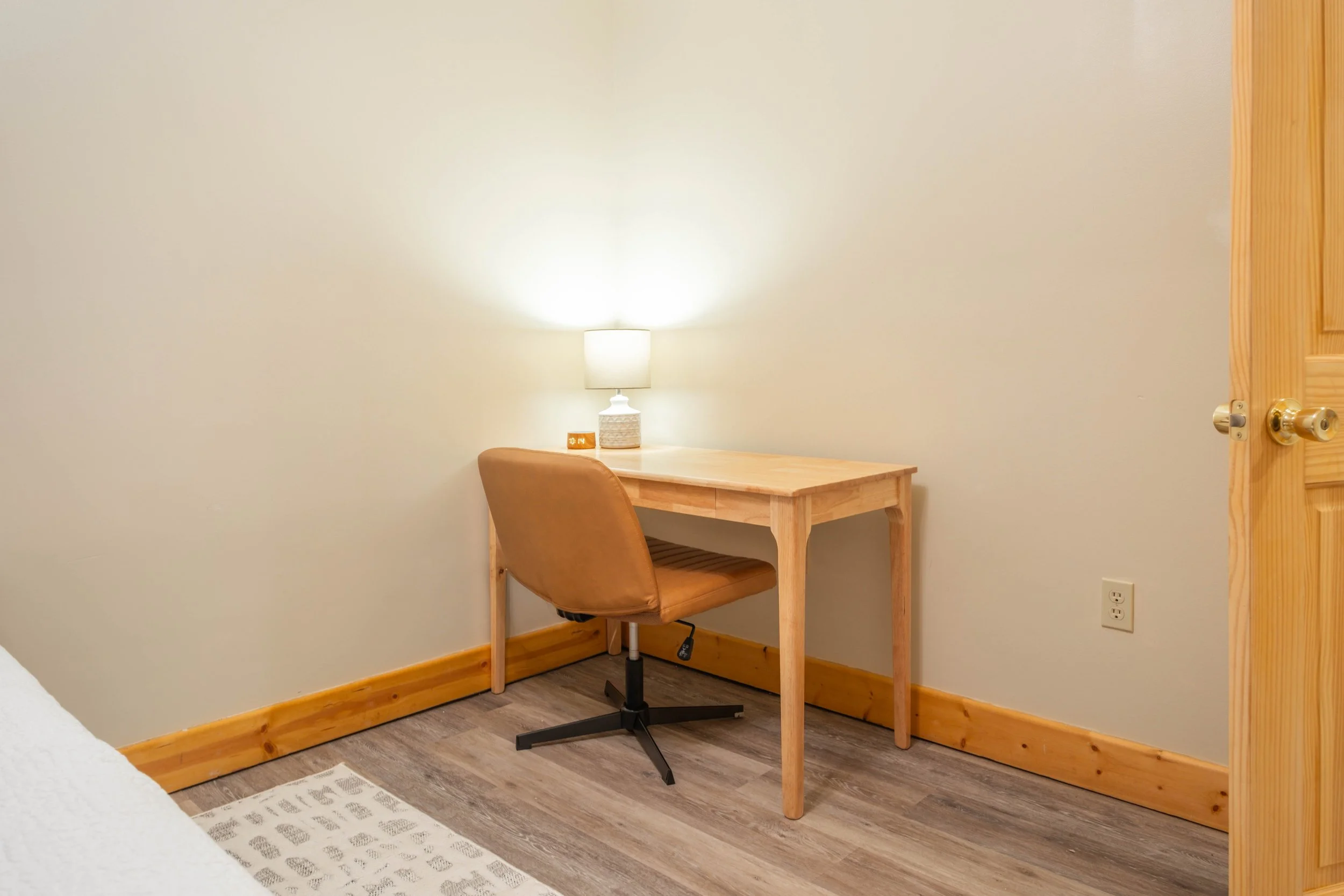 A minimalist bedroom corner with a wooden desk, a brown swivel chair, and a table lamp on the desk, against white walls and wooden baseboards.