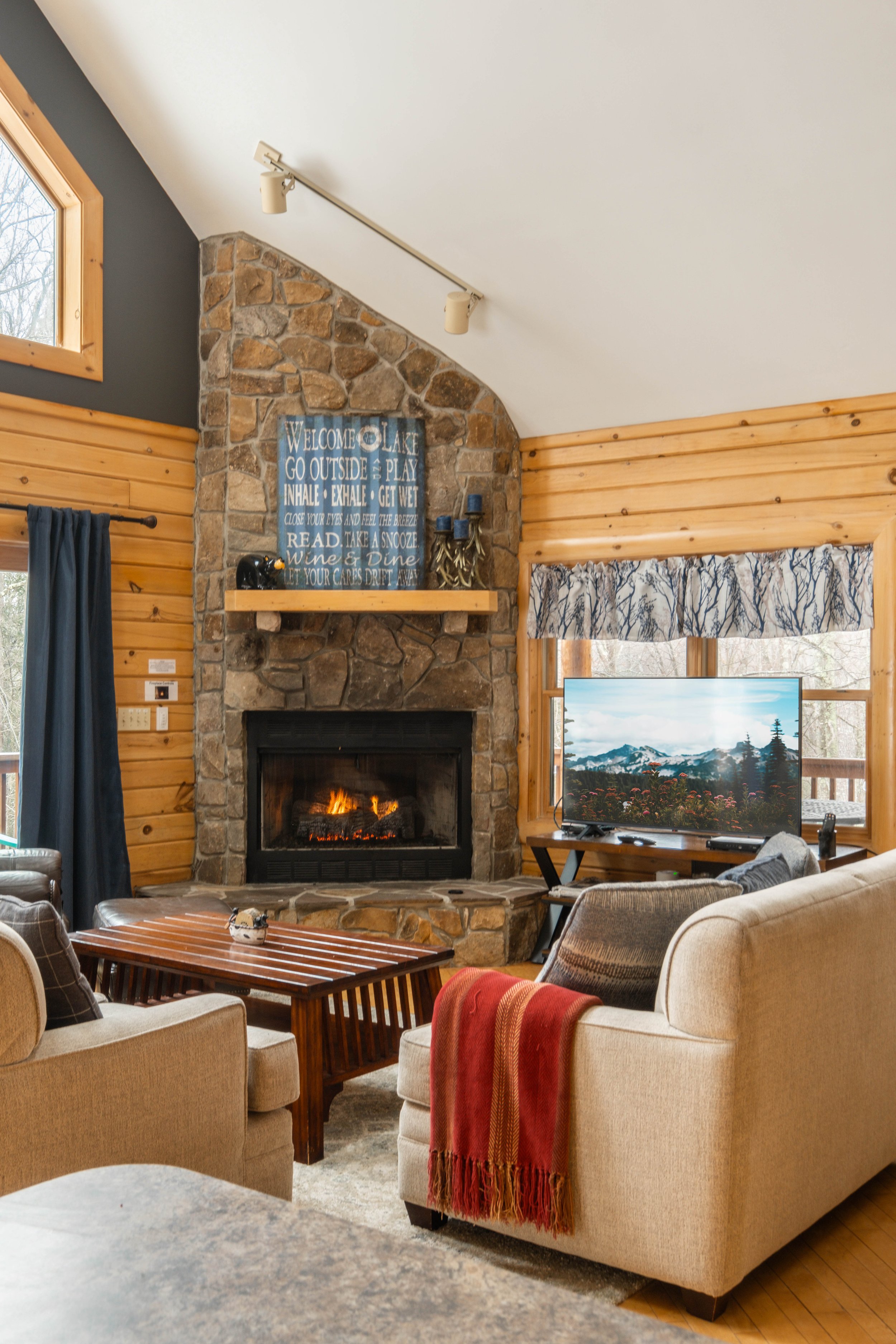 Cozy living room with a stone fireplace, beige couches, wooden coffee table, TV, and wood-paneled walls.