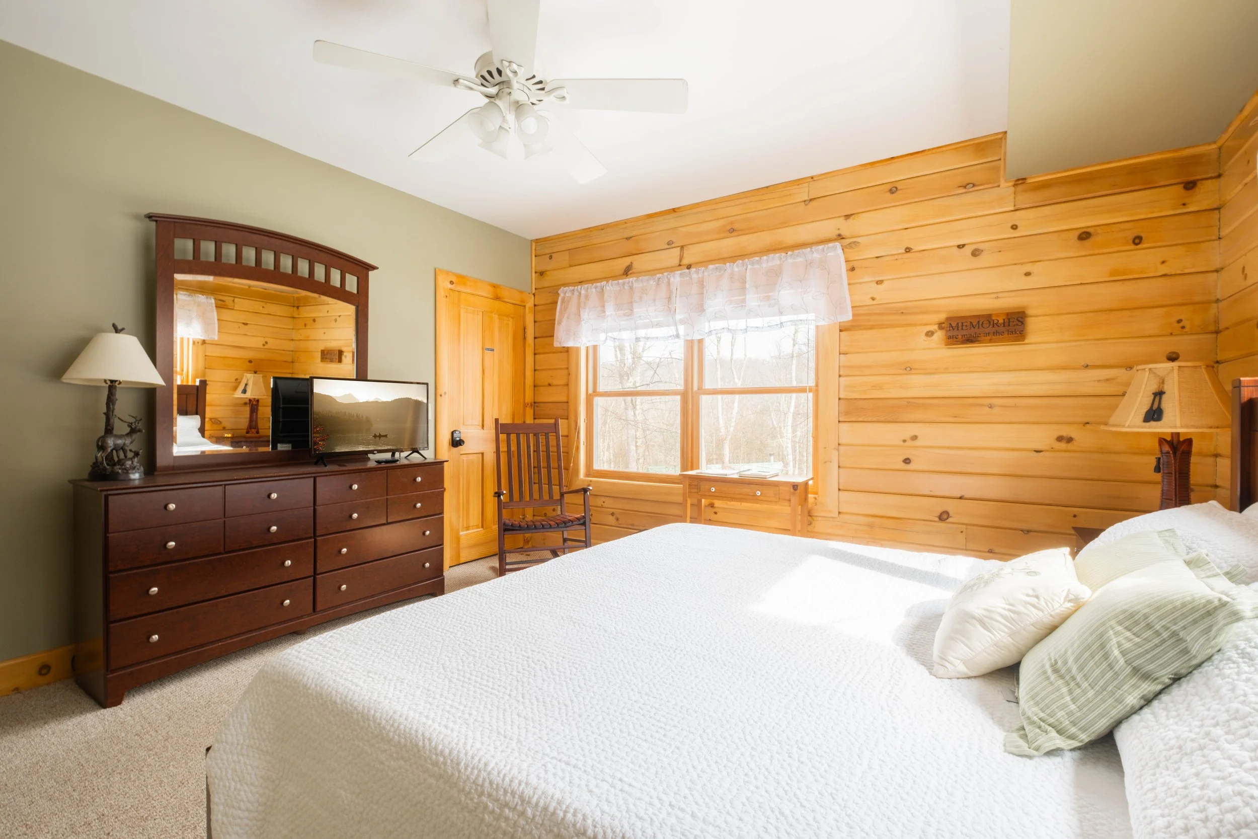 A cozy bedroom with wooden wall paneling, a large bed with pillows, a dark wooden dresser with a mirror and a television, a rocking chair next to a window, and a ceiling fan.