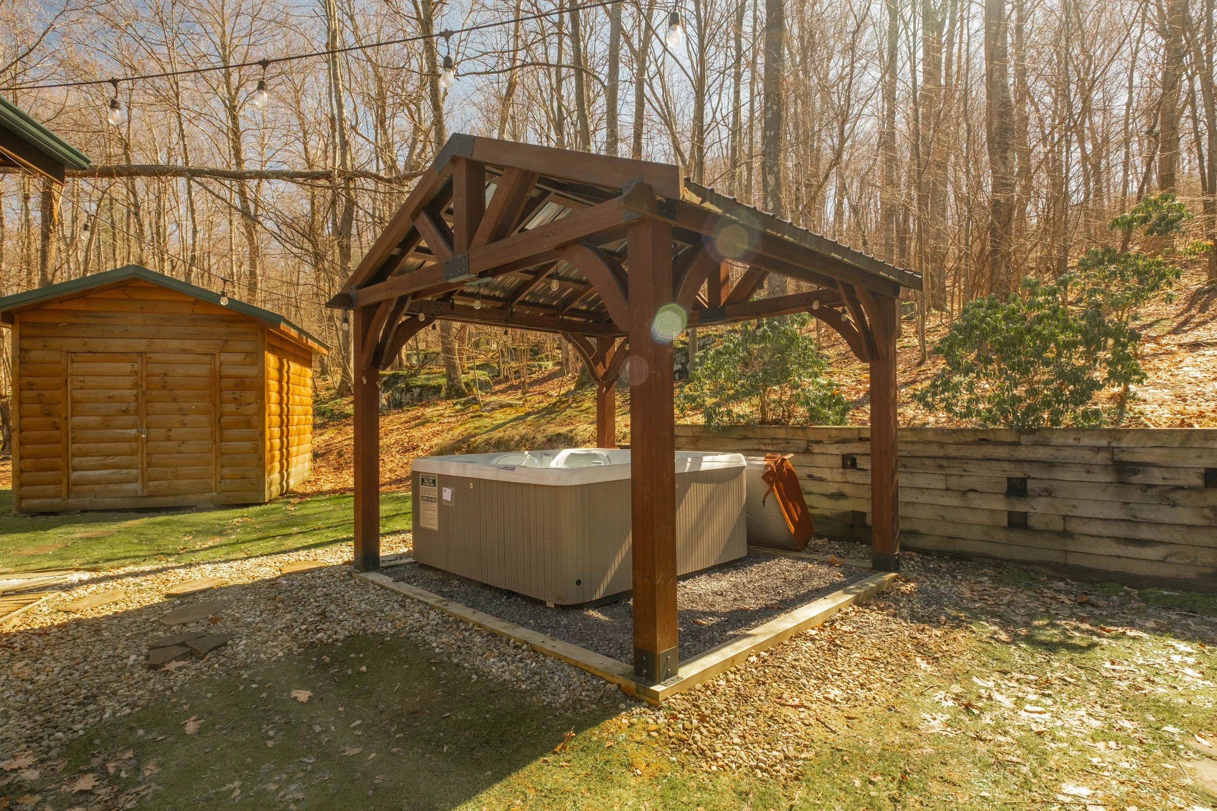 An outdoor hot tub under a wooden shelter in a backyard with a wooded hillside background, a wooden shed, and a retaining wall.