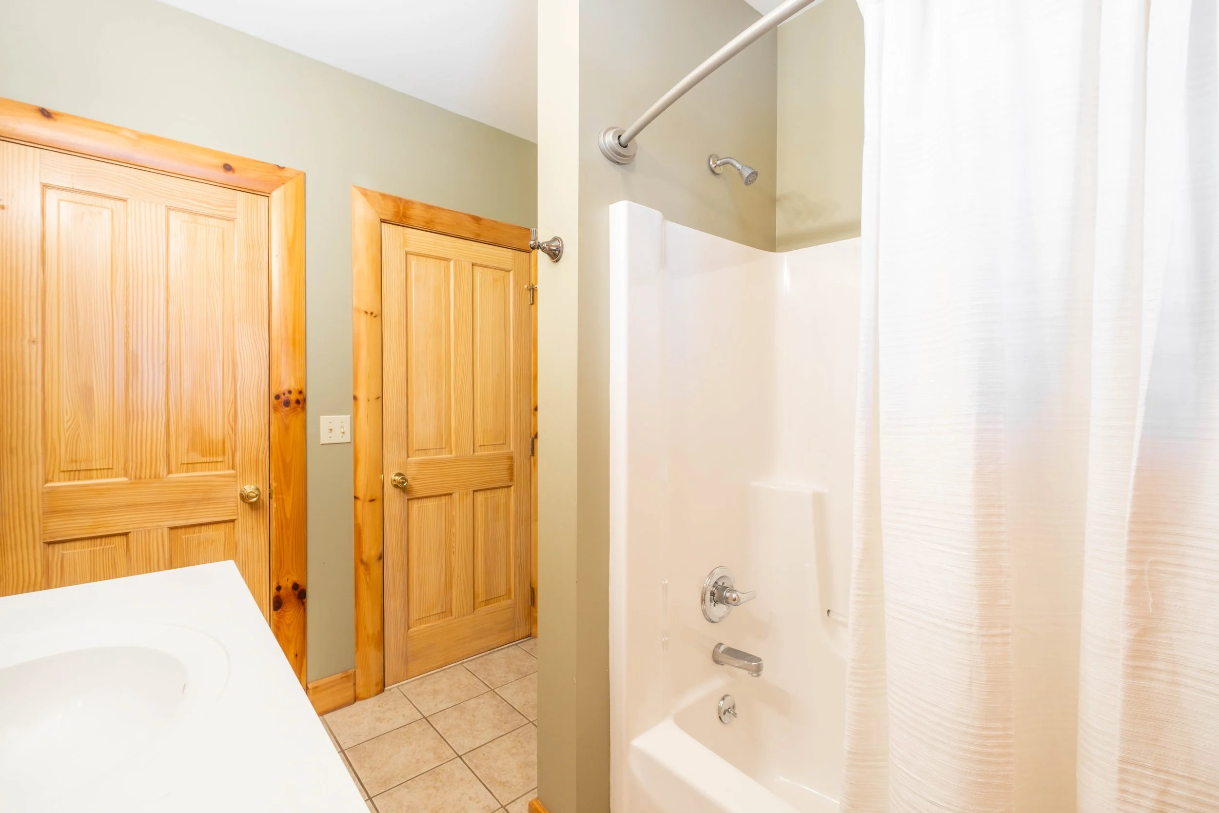 Bathroom with two wooden doors, a light-colored wall, a corner of white countertop with sink, tiled floor, and a shower with a curtain and white fiberglass surround.