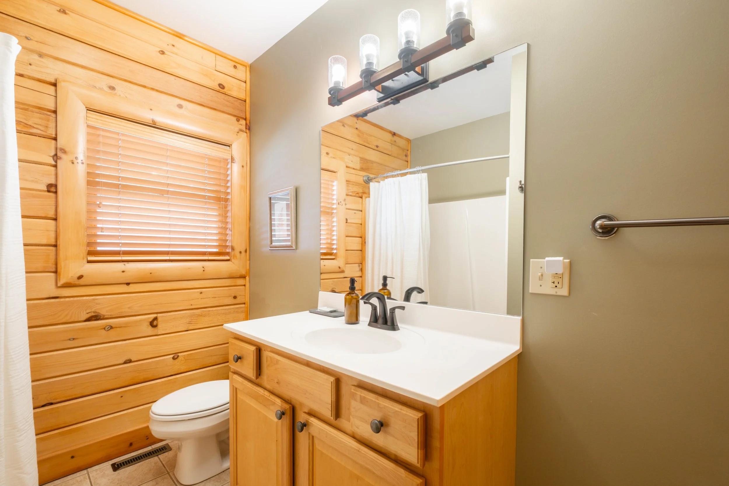 Bathroom with wood-paneled wall, window with blinds, mirror, vanity with sink, soap dispenser, and toothbrush holder, shower curtain, and towel bar.