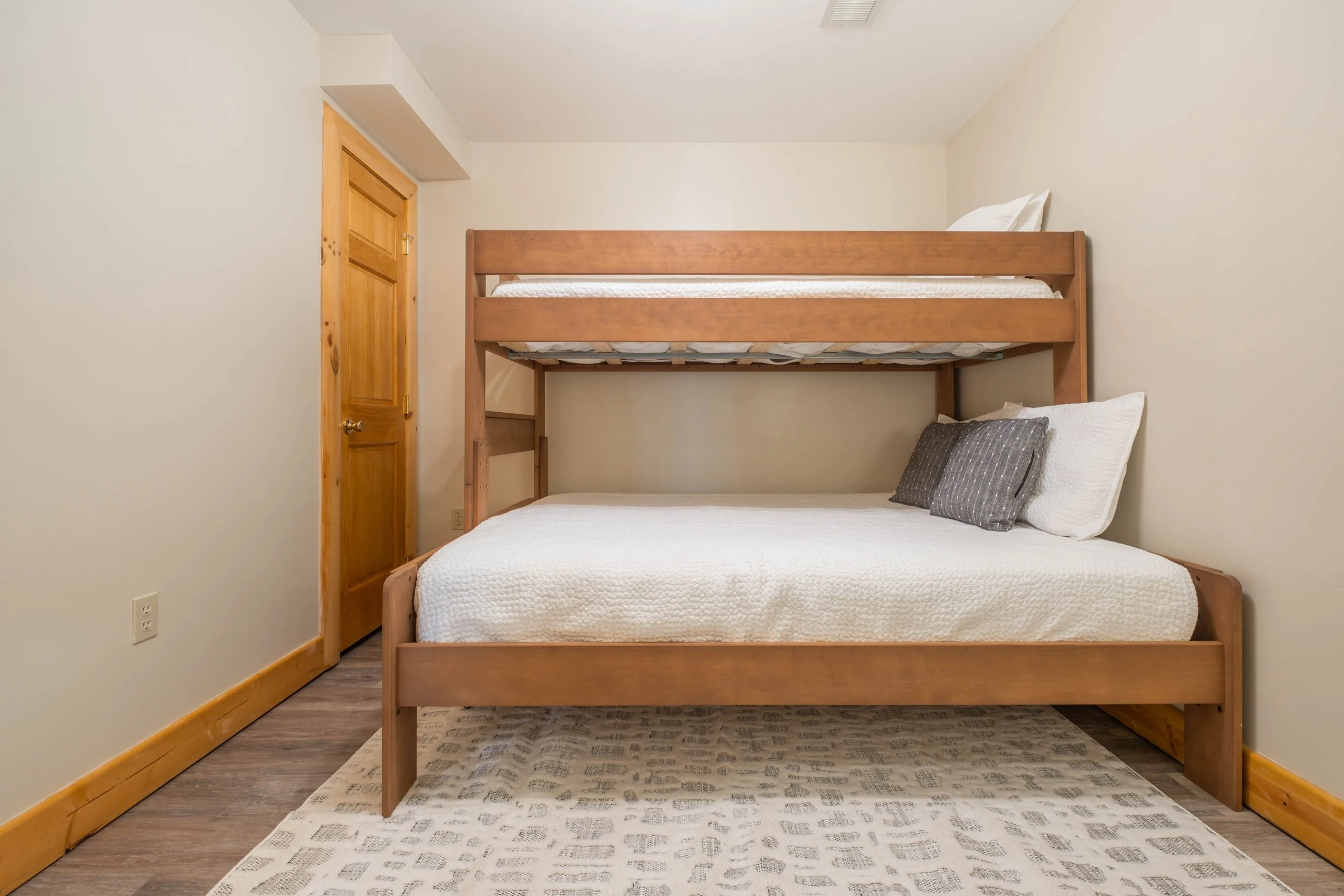 Bedroom with a wooden bunk bed, white bedspread, gray pillows, wooden door, and a cream-colored rug.