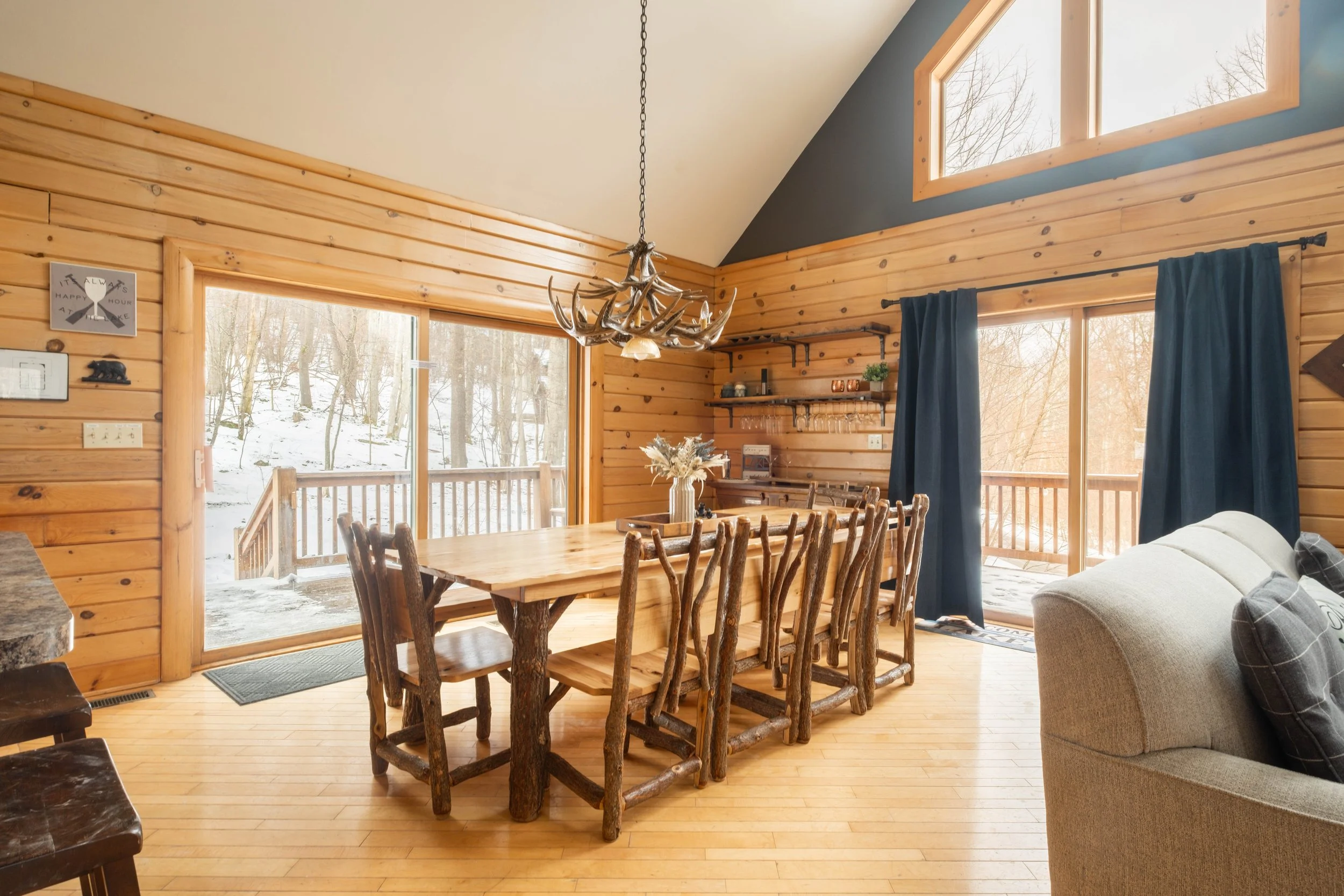 Interior of a cozy cabin dining area with a large wooden table, rustic chairs, and scenic snow-covered view outside through large windows.