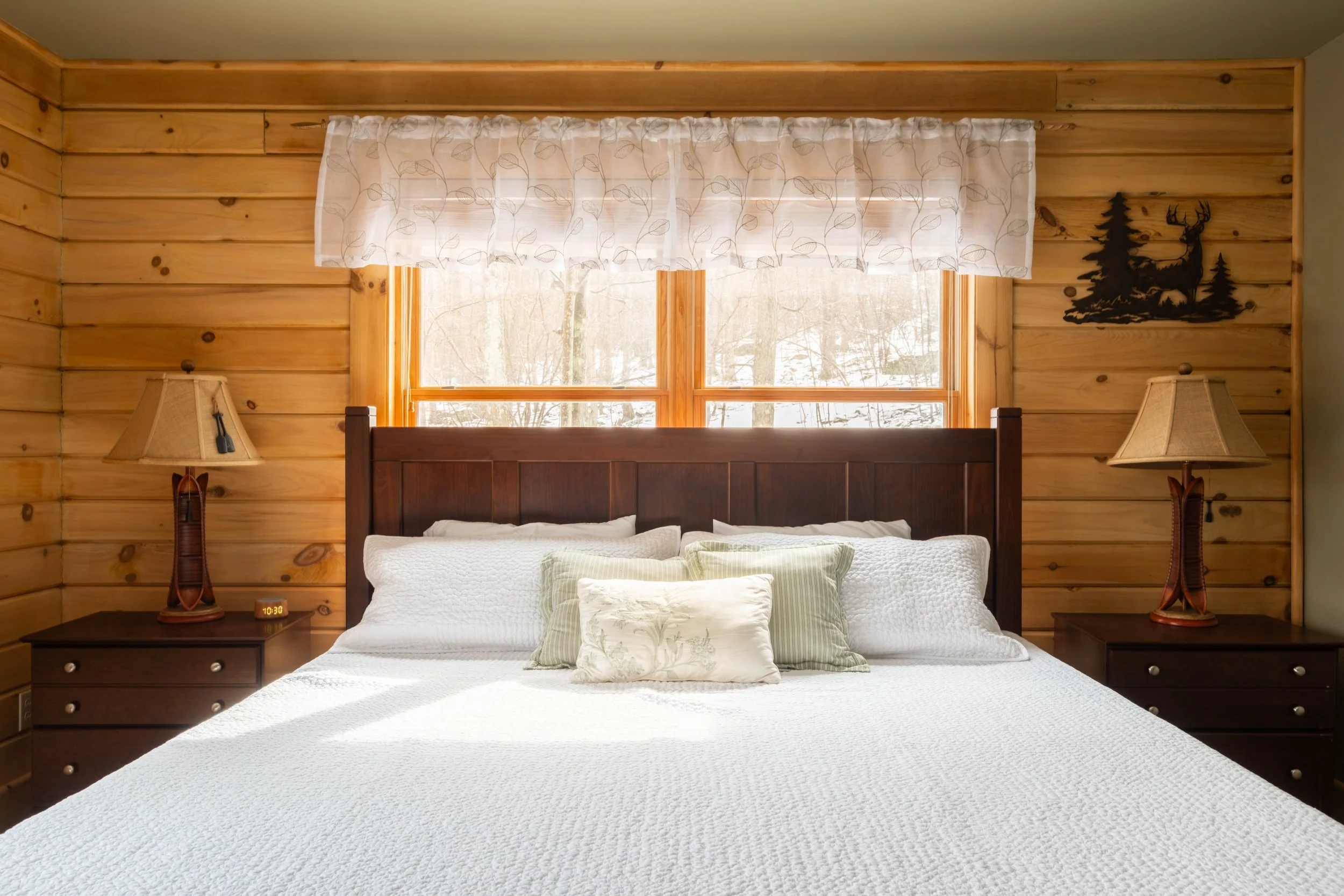 A cozy bedroom with wood-paneled walls, a large bed with white bedding and multiple pillows, two matching wooden nightstands with lamps, and a window with sheer curtains showing snow outside.
