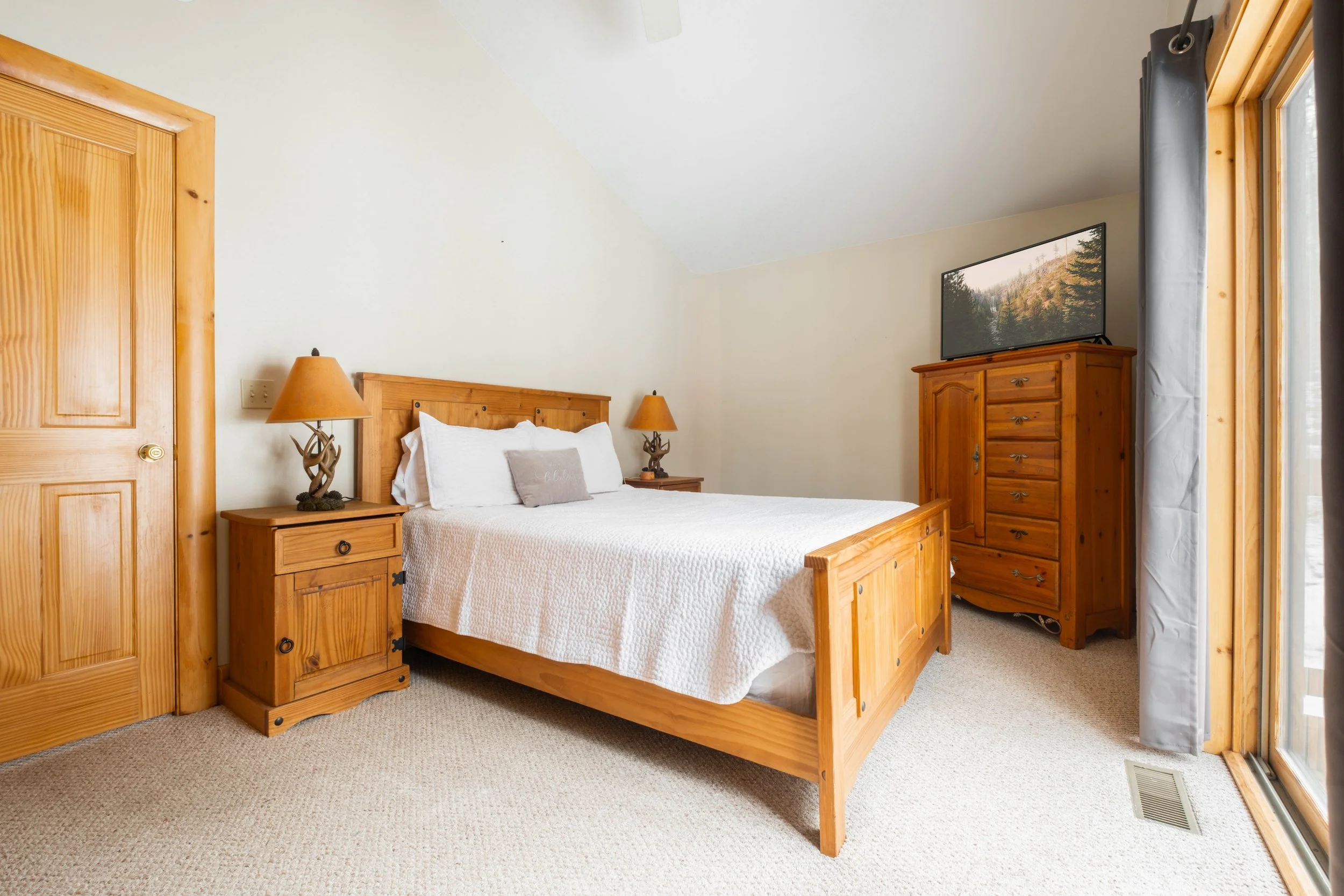 A bedroom with a wooden bed, nightstands, dresser, and a television, with natural light coming in through a sliding glass door with curtains.