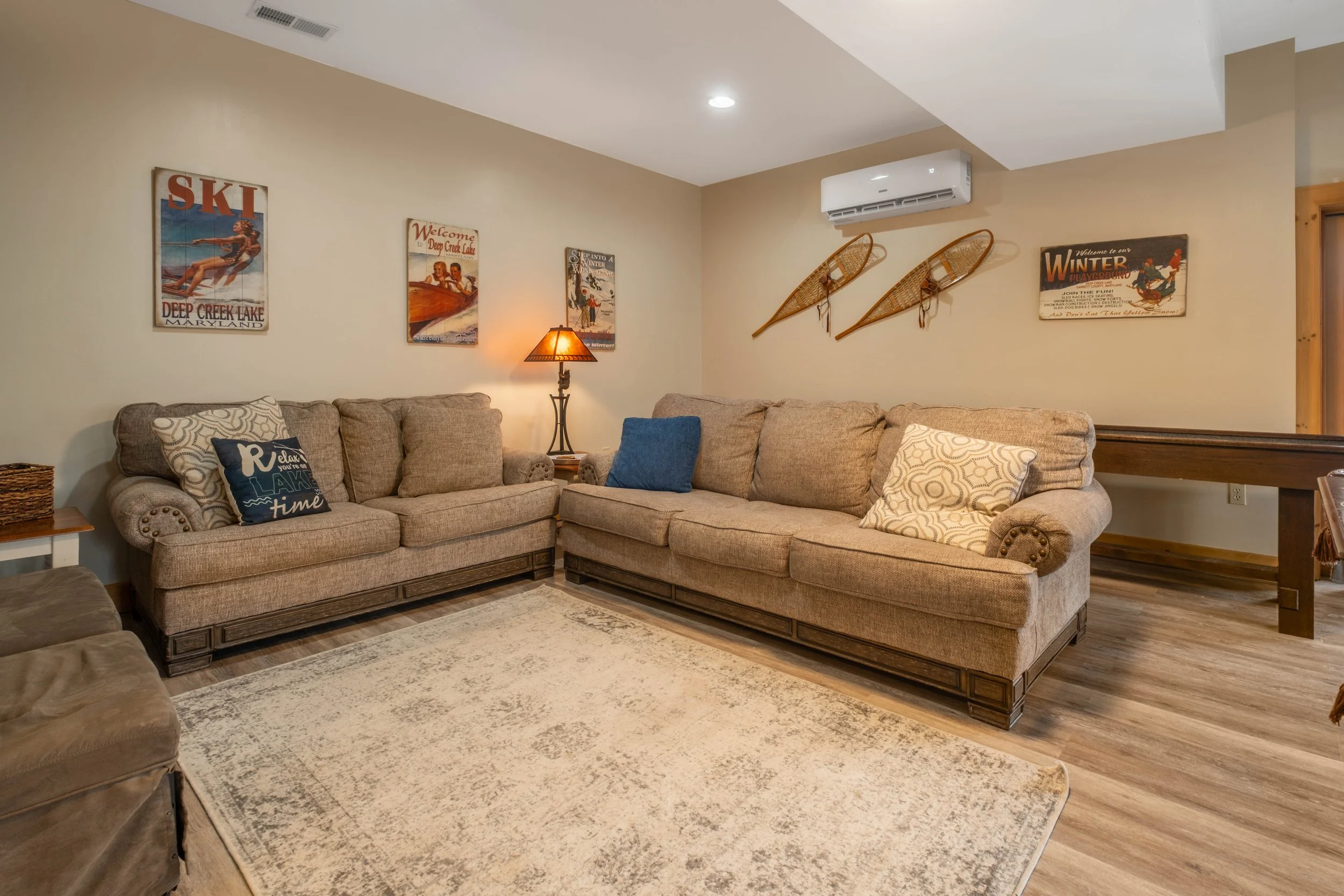 Living room with beige sofas, decorative pillows, a floor lamp, wall art including vintage ski and winter posters, and a mounted air conditioning unit.