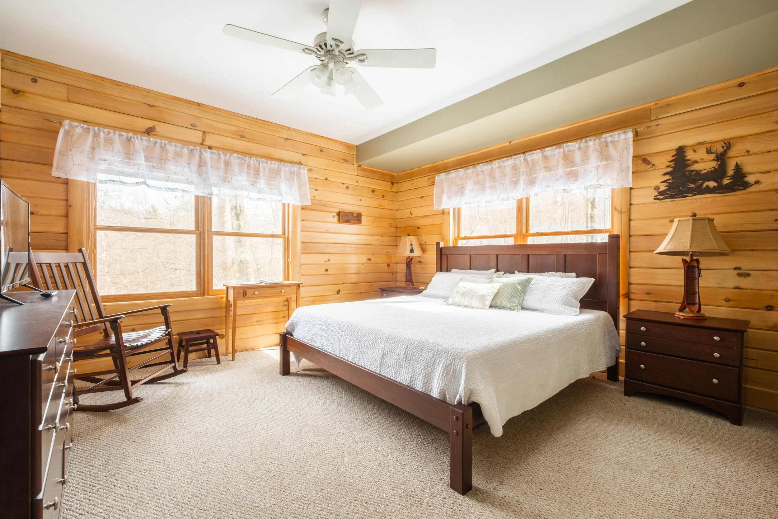 A bedroom with wooden walls, two large windows with curtains, a wooden bed with white bedding, a bedside table with a lamp, a wooden armchair, a small wooden stool, a lamp, a dresser with a TV, and a ceiling fan.