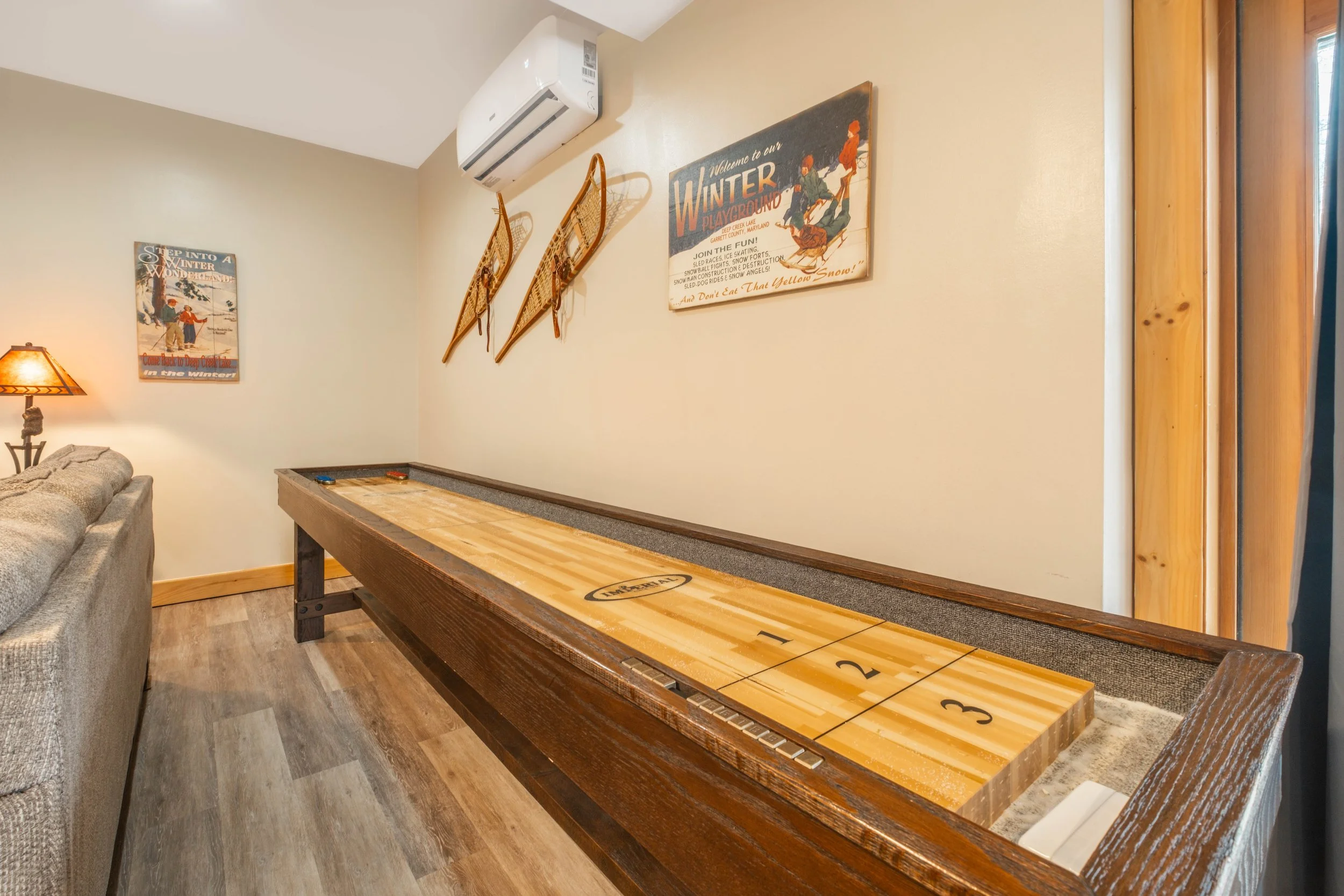 Indoor shuffleboard table with scoring area, decorated with winter-themed vintage posters and sled decor on beige walls, next to a gray sofa and a lamp.