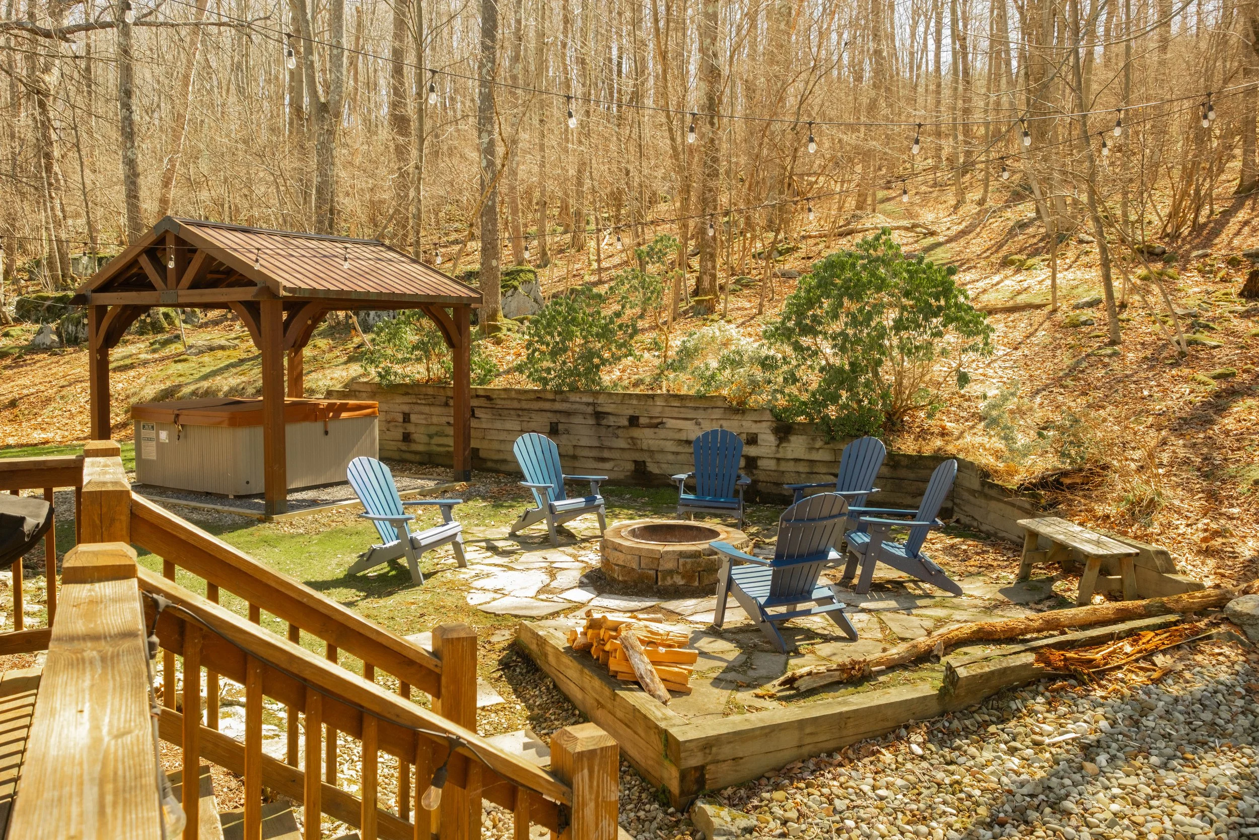 Wooden outdoor firepit with six blue Adirondack chairs arranged around it, located in a wooded backyard with a small hot tub and string lights overhead.