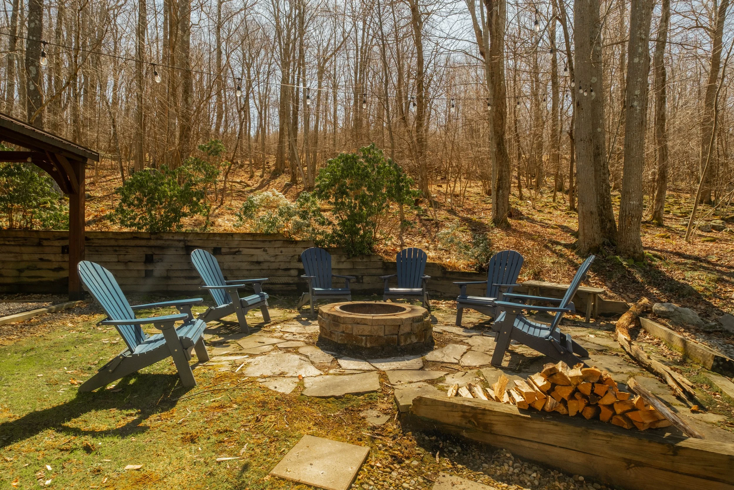 Six Adirondack chairs arranged in a circle around a brick fire pit in a backyard with fallen leaves, firewood, and a wooded hillside in the background.