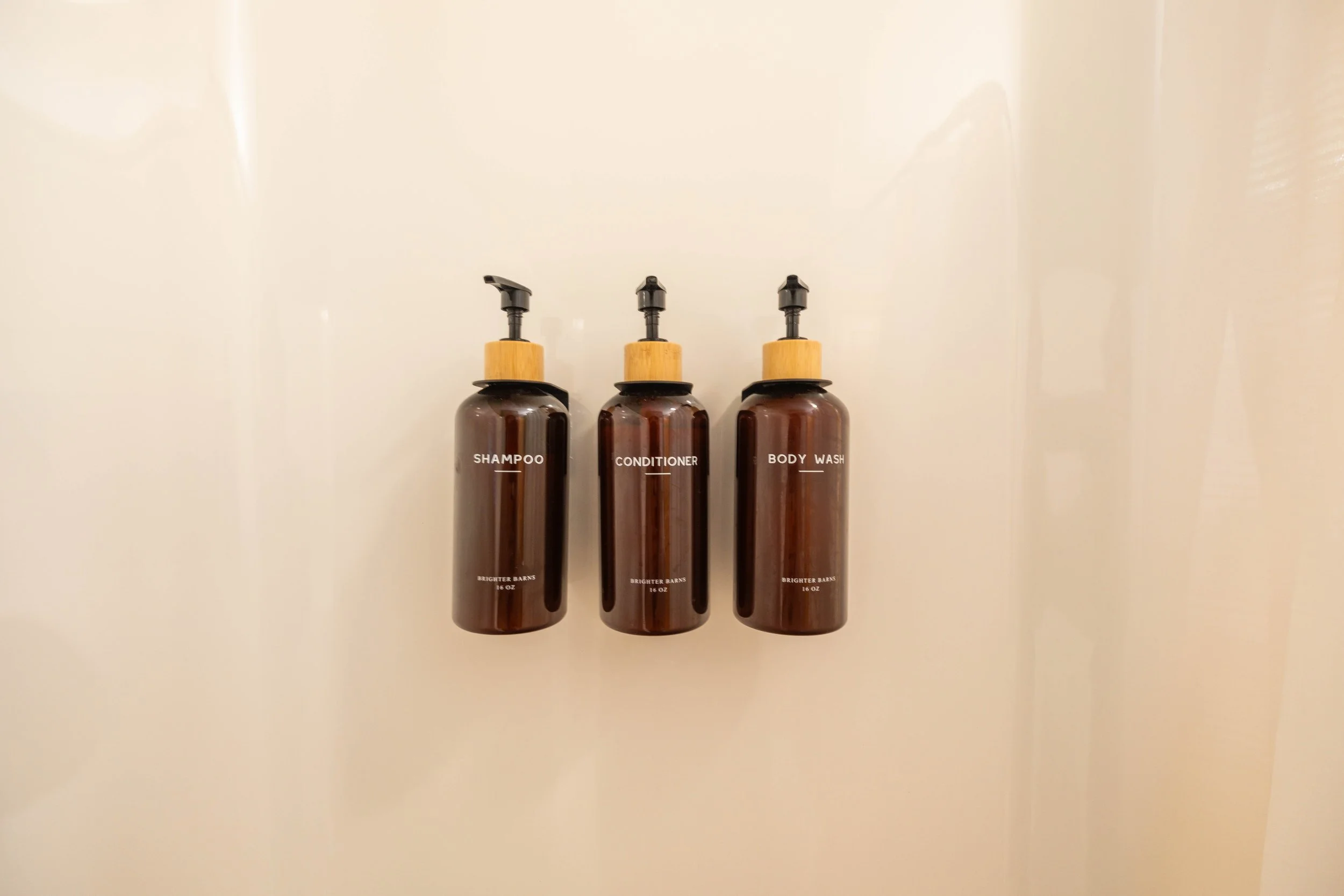 Three brown bottles labeled shampoo, conditioner, and body wash with black pump tops, mounted on a cream-colored wall.