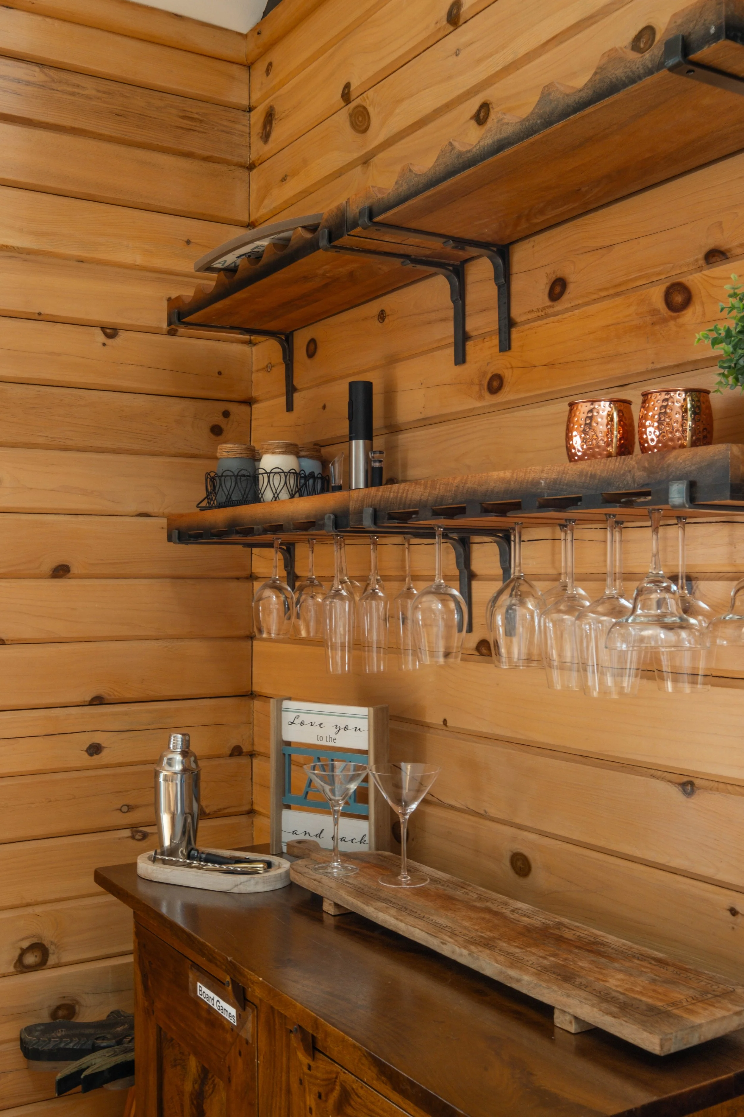 Wooden bar area with two shelves holding glassware, cups, and decorative items. A wooden countertop with a tray and cocktail shaker, with a framed sign and a pair of empty martini glasses.