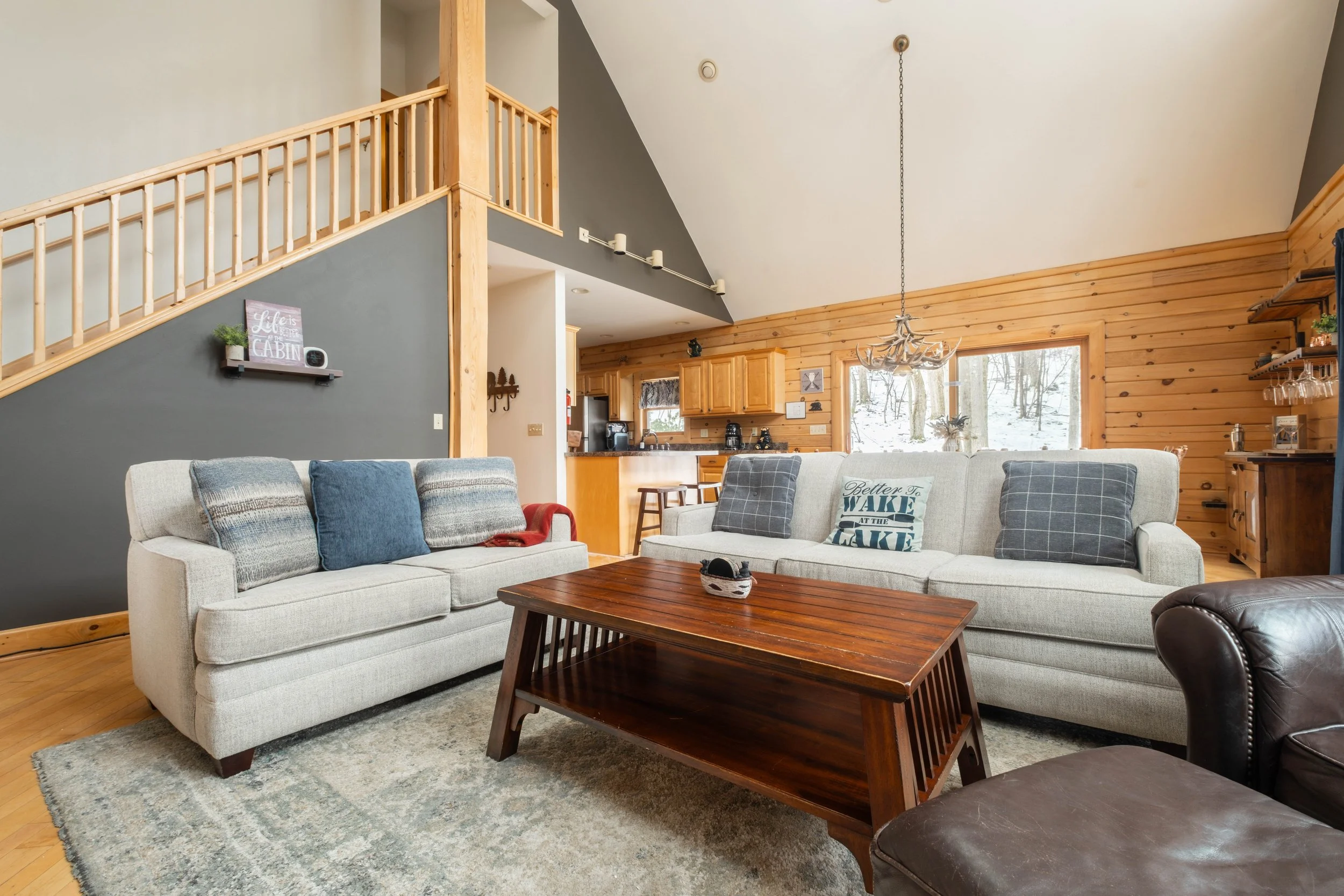 Living room with beige couches, a wooden coffee table, and a leather armchair, with wood-paneled walls, a staircase, and a kitchen area in the background.