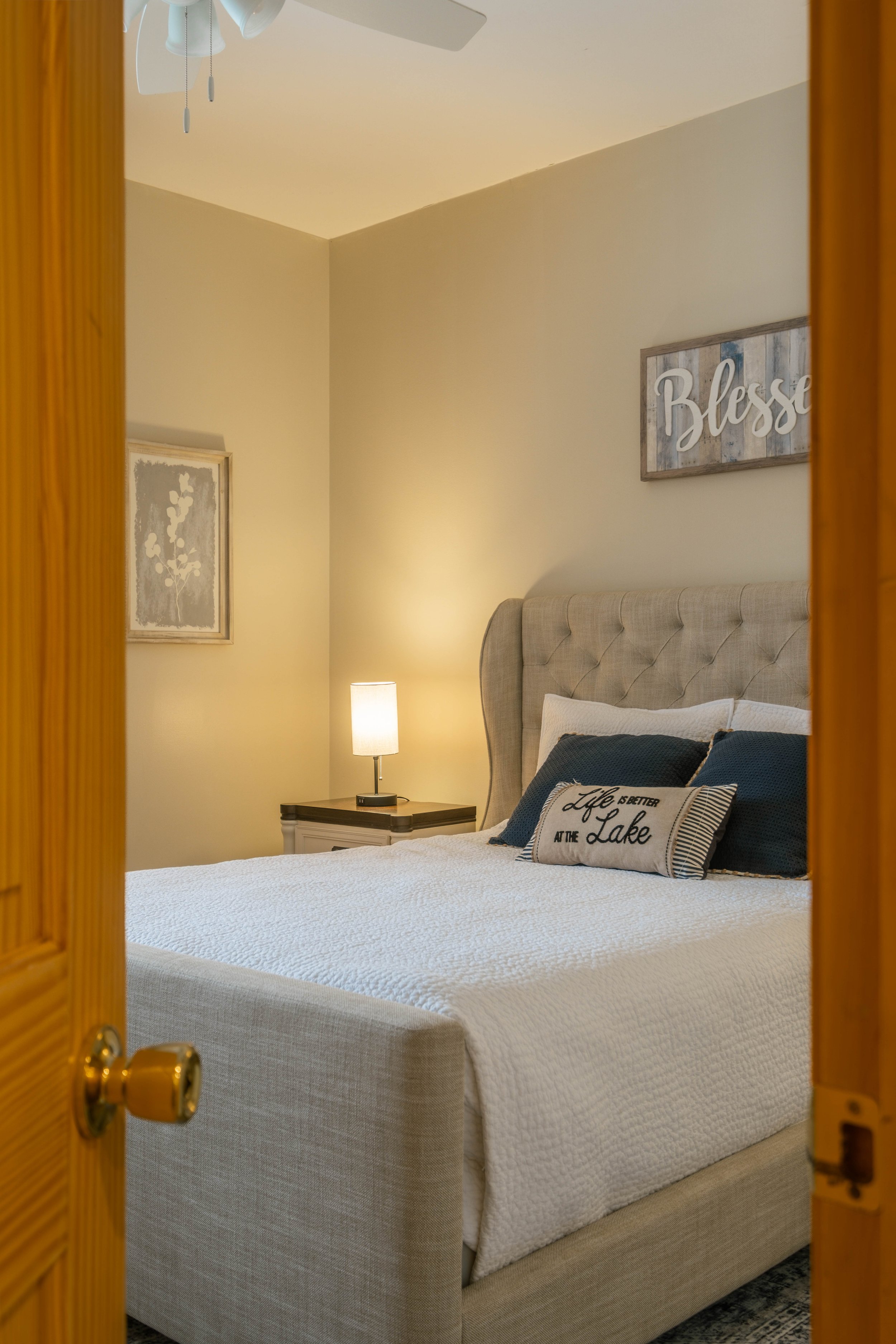 A cozy bedroom seen through a partially open wooden door, featuring a beige upholstered bed with decorative pillows, a nightstand with a lamp, and wall art with the words 'Blessed' and 'Life is better at the Lake'.