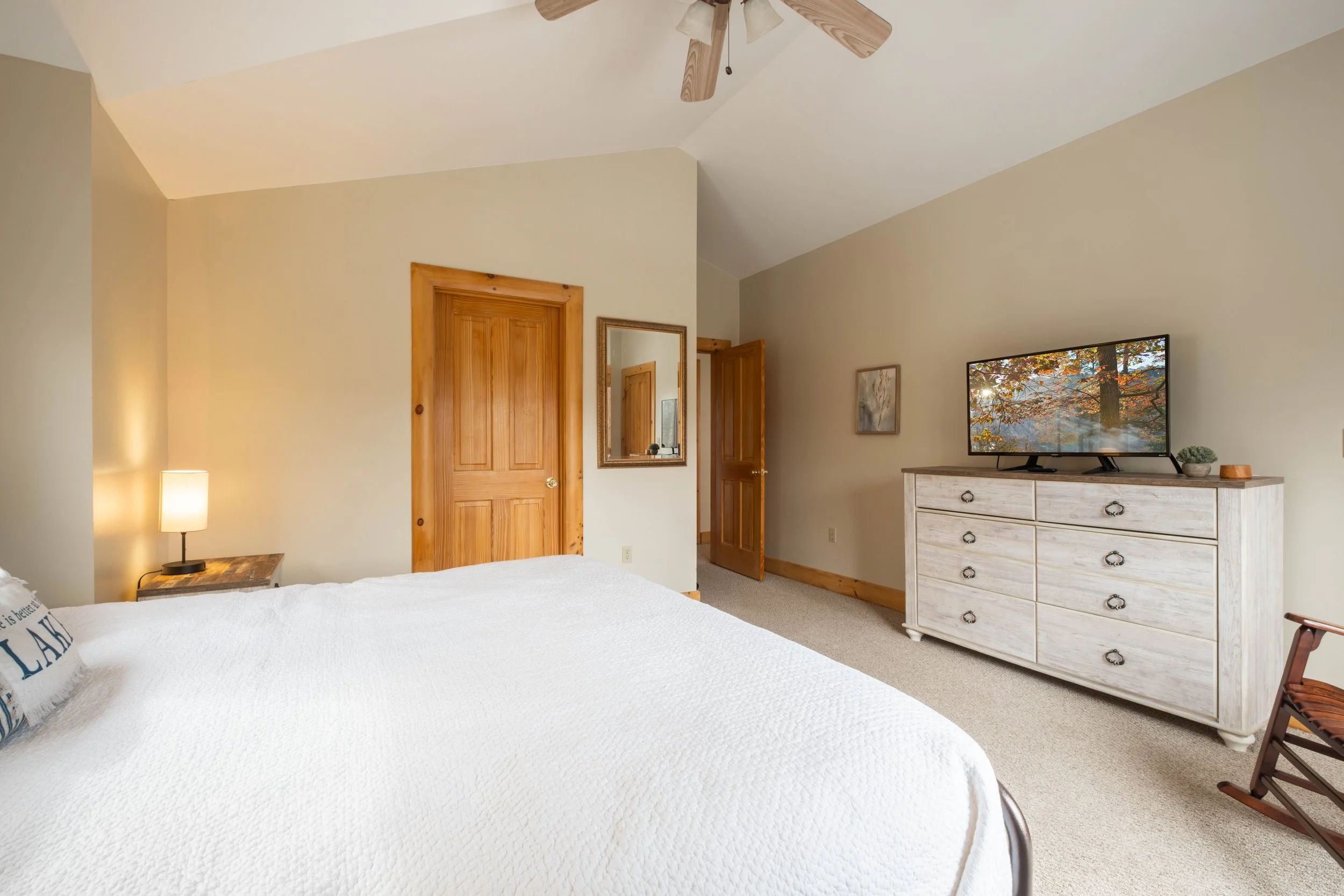 A bedroom with a white bed, wooden door, a dresser with a television, wall art, a mirror, and a ceiling fan.