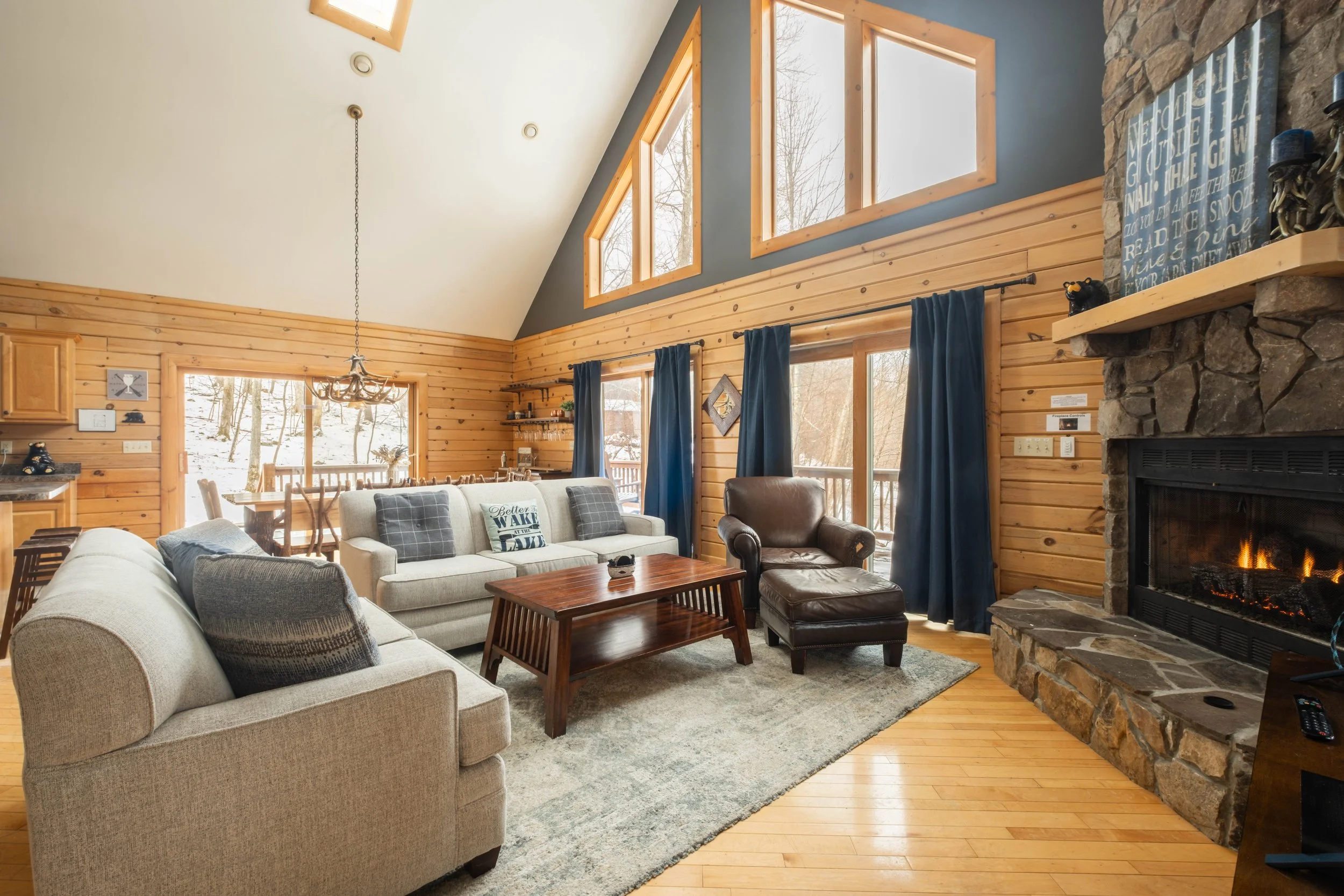 Cozy living room with wood-paneled walls, large windows, a stone fireplace with a burning fire, beige and brown sofas, a wooden coffee table, and blue curtains, in a rustic cabin style.