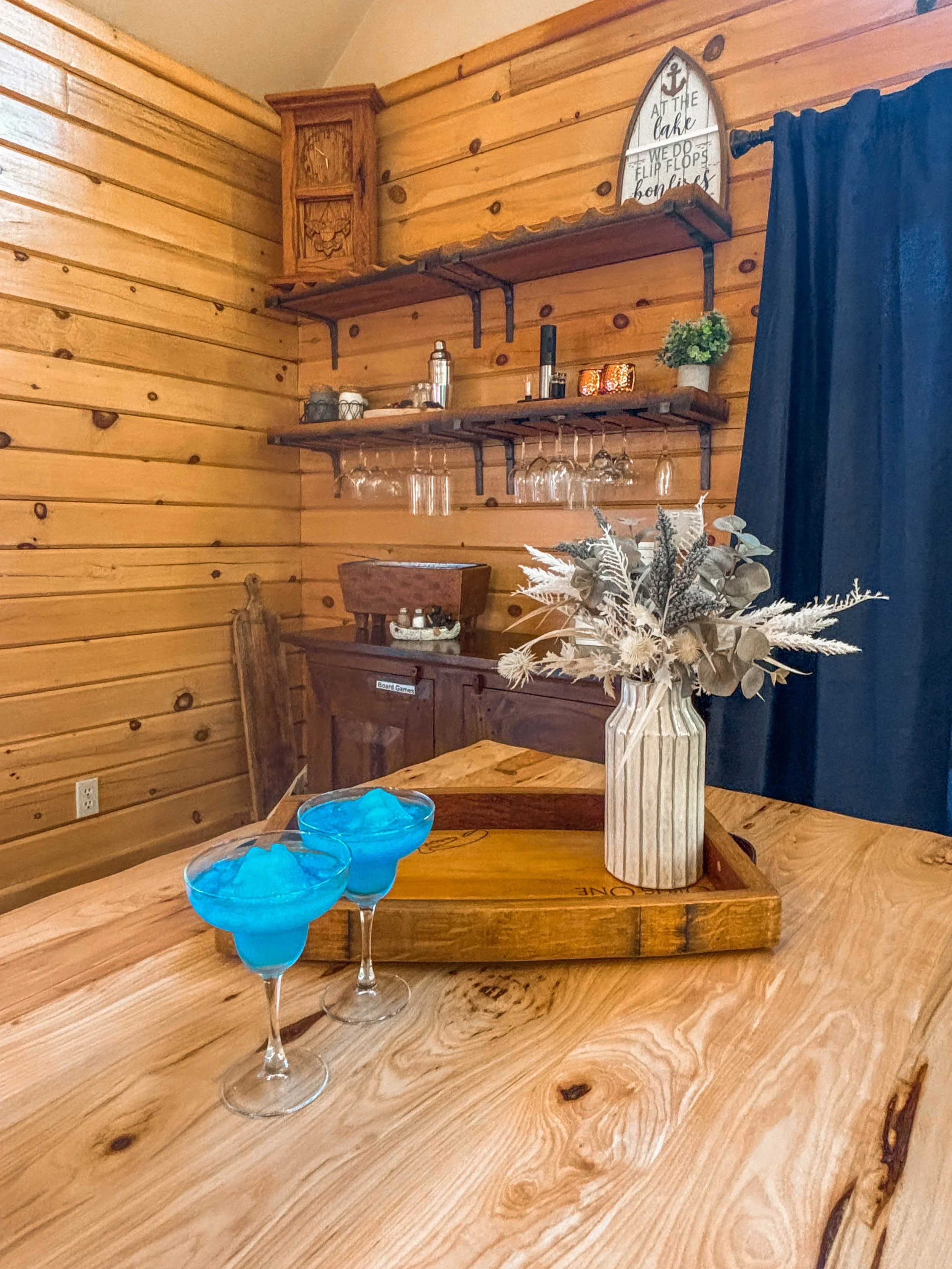 A wooden dining table with two blue cocktails, a striped vase with dried flowers, and a tray, set against a background of wooden walls and shelves holding wine glasses and decor, with a dark blue curtain on the right.