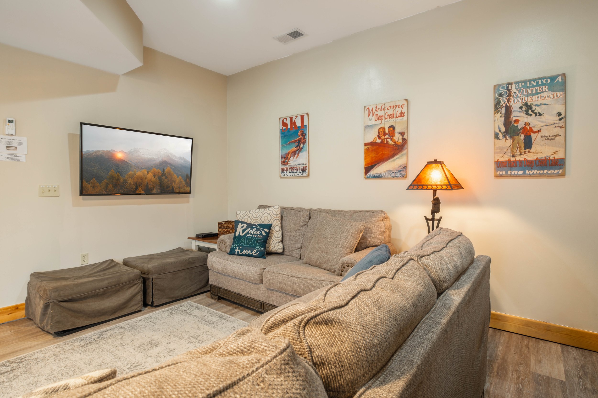 Living room with beige walls, a wall-mounted flat-screen TV displaying mountains, a beige couch with pillows, a floor lamp, and framed vintage winter sports posters on the wall.