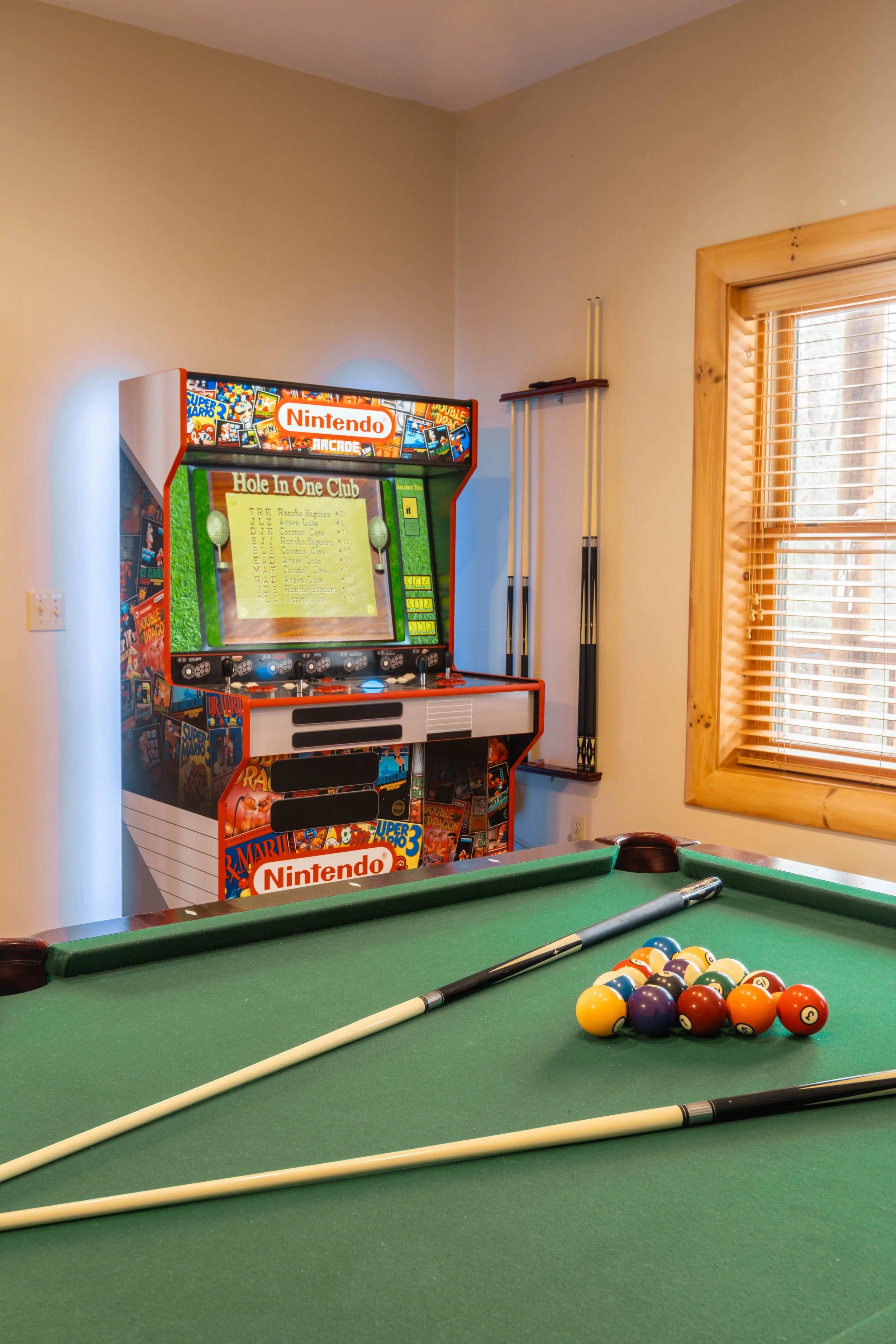 A pool table with balls racked in a triangle and two cues on the green felt surface, with a vintage Nintendo arcade game in the background beside a window with wooden blinds.