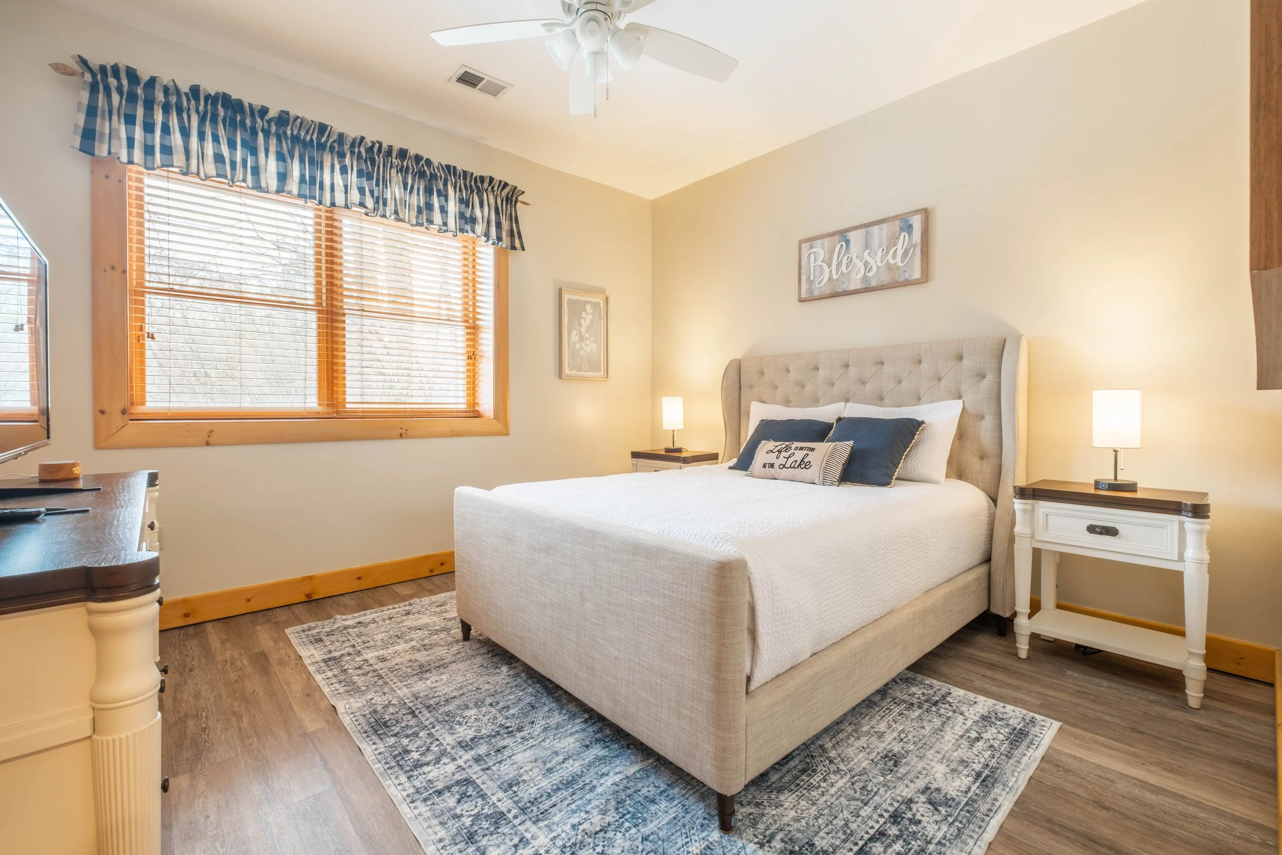 A cozy bedroom with a large bed that has a tufted headboard, flanked by two nightstands with lamps. The room has a large window with wooden blinds and checkered valance, and light-colored walls decorated with artwork and a sign that says "Blessed." T