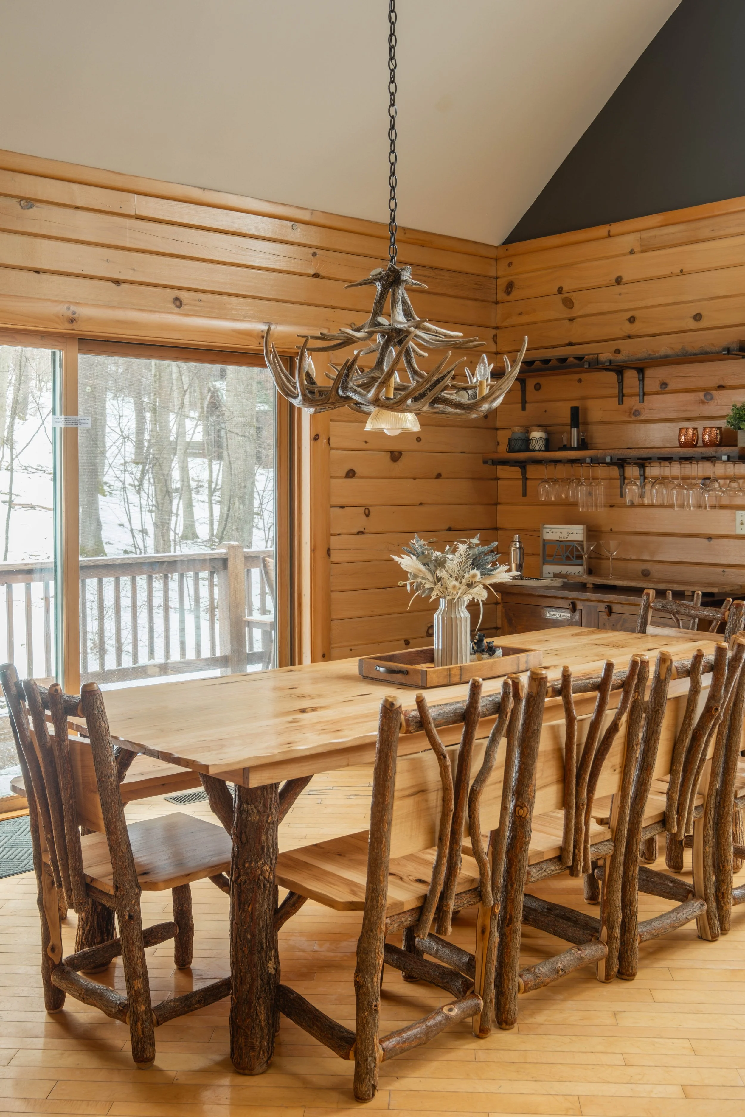 Rustic dining area with a wooden table, matching chairs, a chandelier made of antlers, and shelves with glasses and decor, in a cabin-style room with wood-paneled walls and a sliding glass door leading to a snowy outdoor deck.