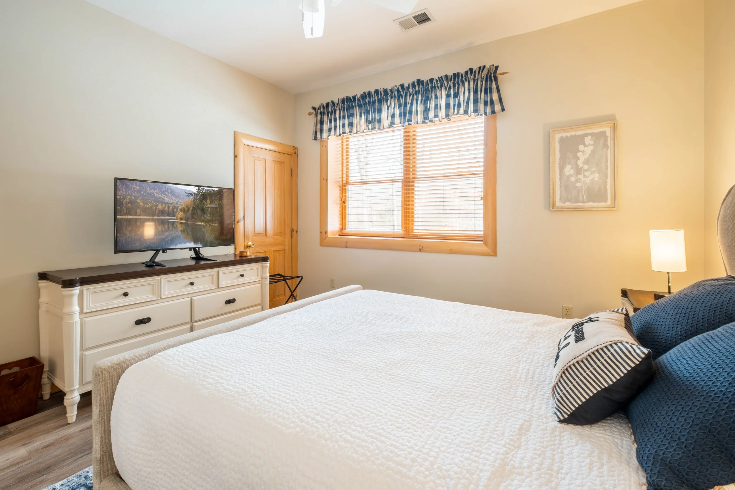 A cozy bedroom with a large bed covered in a white quilt, blue pillows, a wooden nightstand with a lamp, a dresser with a television, a window with wooden blinds and a blue plaid valance, light-colored walls, and a framed artwork on the wall.