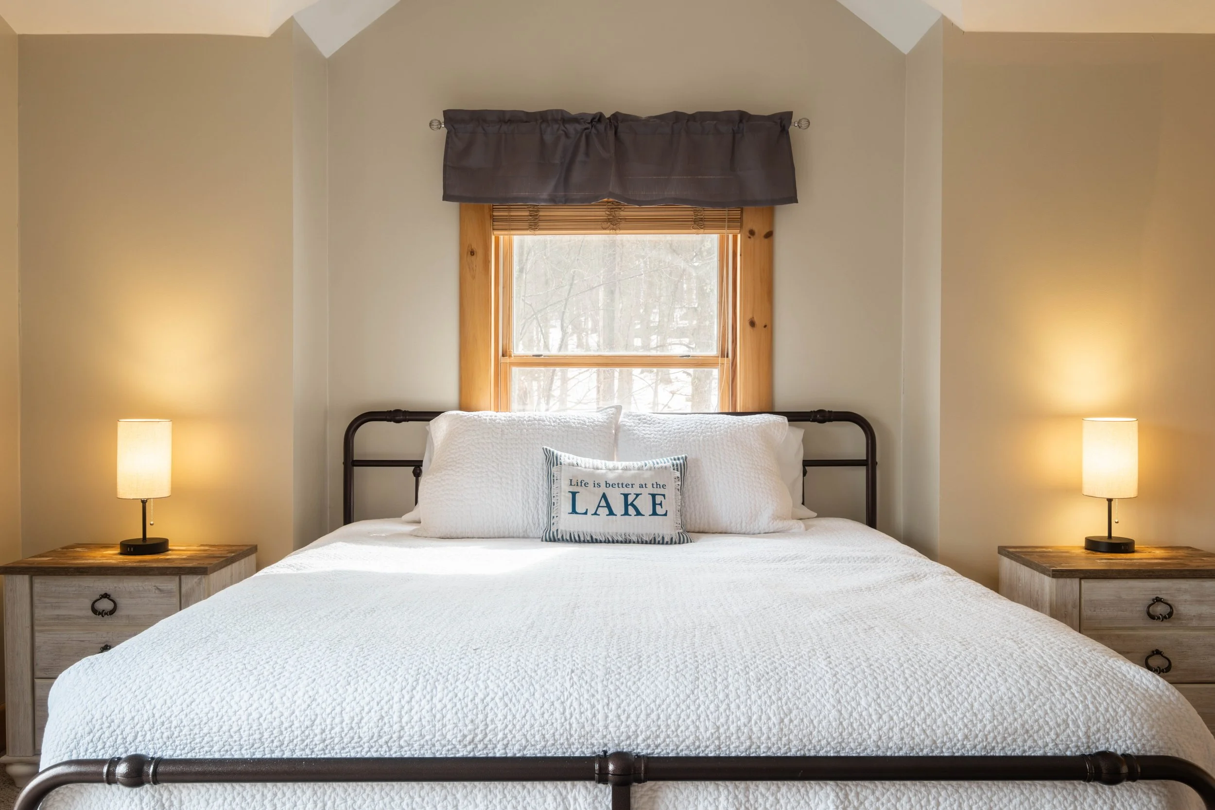 A cozy bedroom with a king-size bed, white bedding, and decorative pillows, one of which reads 'Life is better at the LAKE.' There are two matching nightstands with table lamps on each side of the bed, and a window with wooden trim and a small curtai