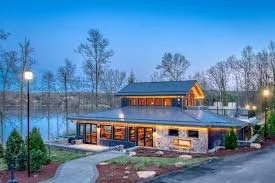 A lakeside lodge or restaurant with large windows, surrounded by trees, with outdoor lights illuminating the area during dusk or early evening.