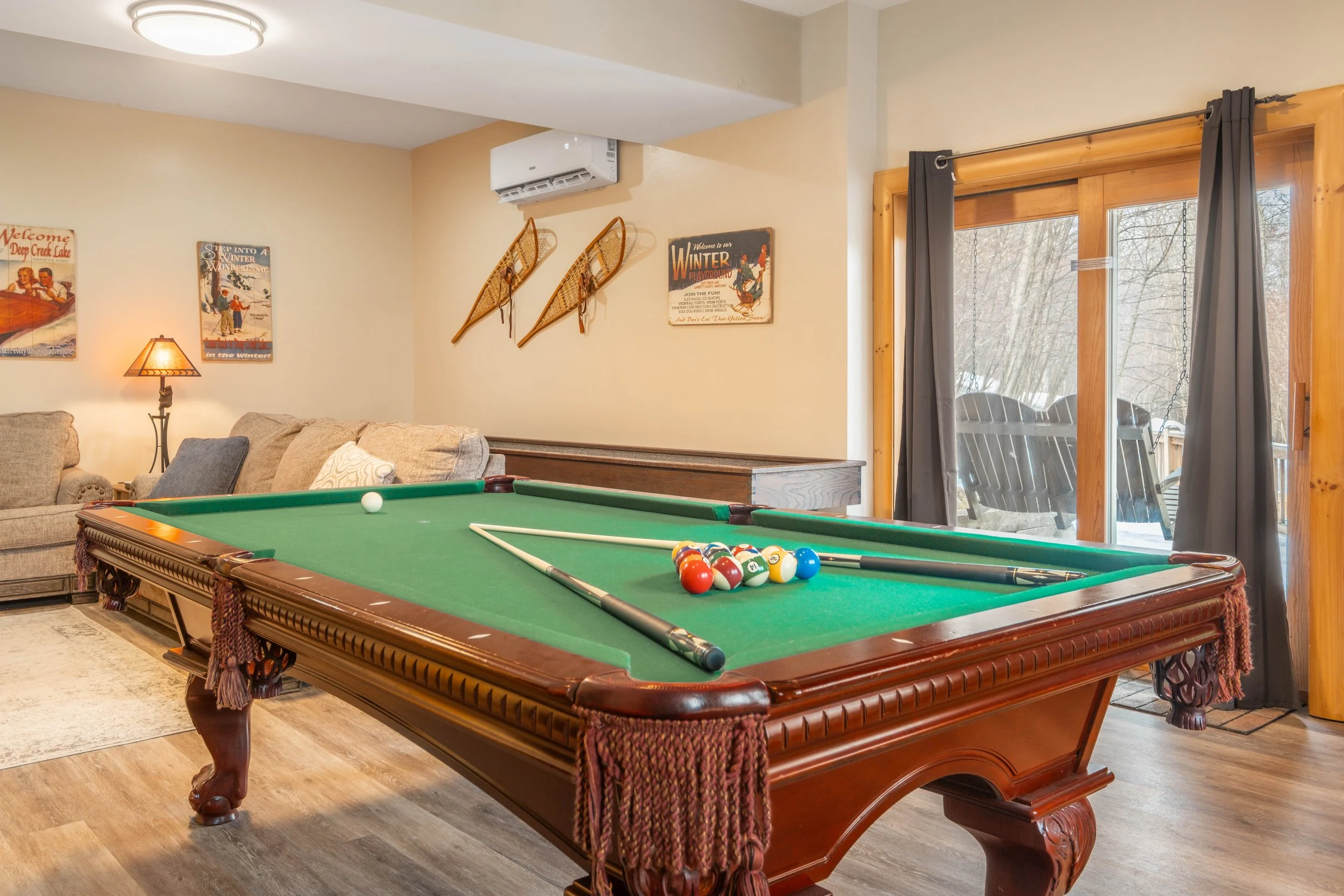 Indoor living room with a pool table, framed posters, snowshoes on the wall, a sofa, a lamp, and a sliding glass door leading outside to a porch with chairs.