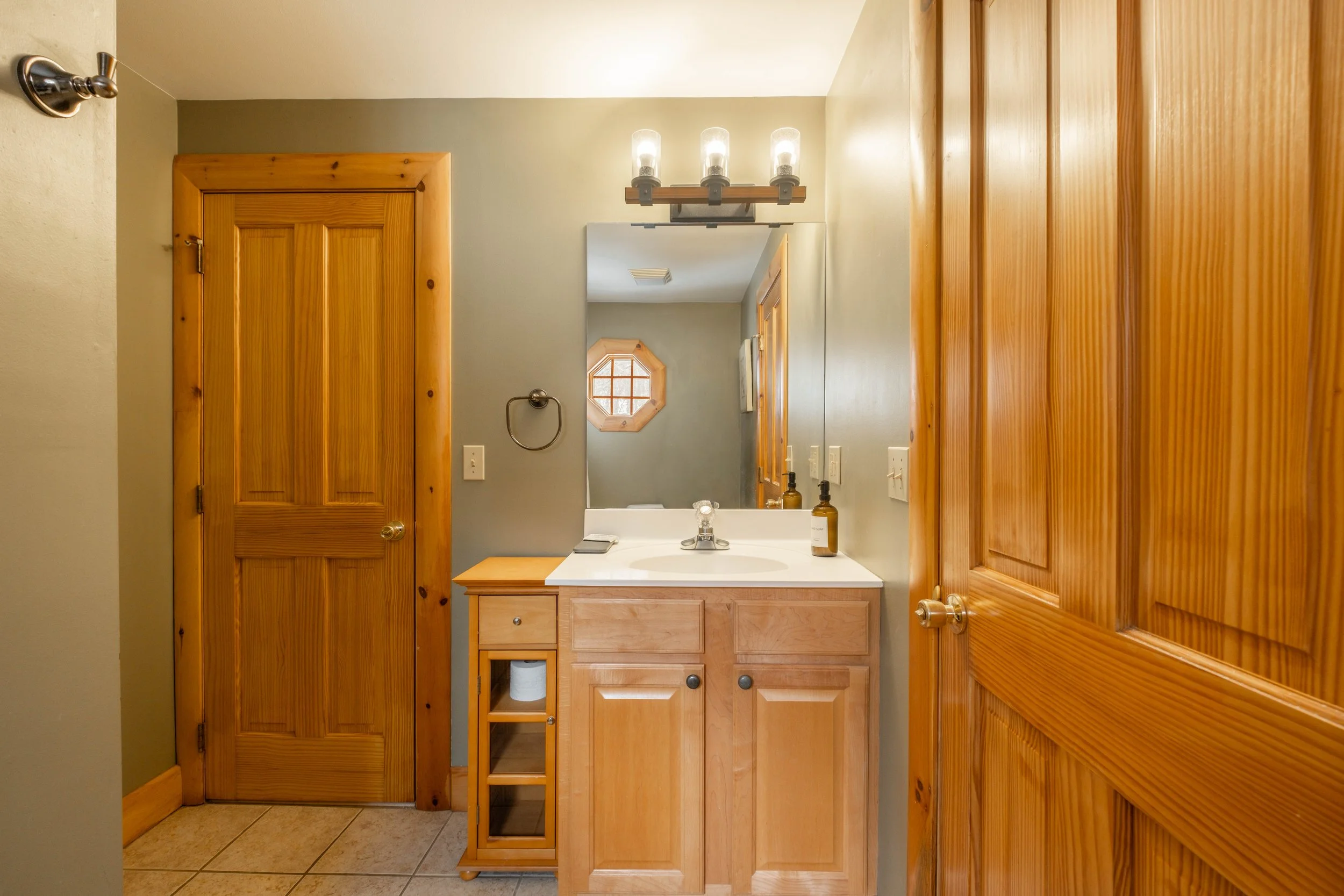 Bathroom with wooden doors, a small wooden cabinet, a mirror, a sink with soap and a bottle, and a wall-mounted light fixture.