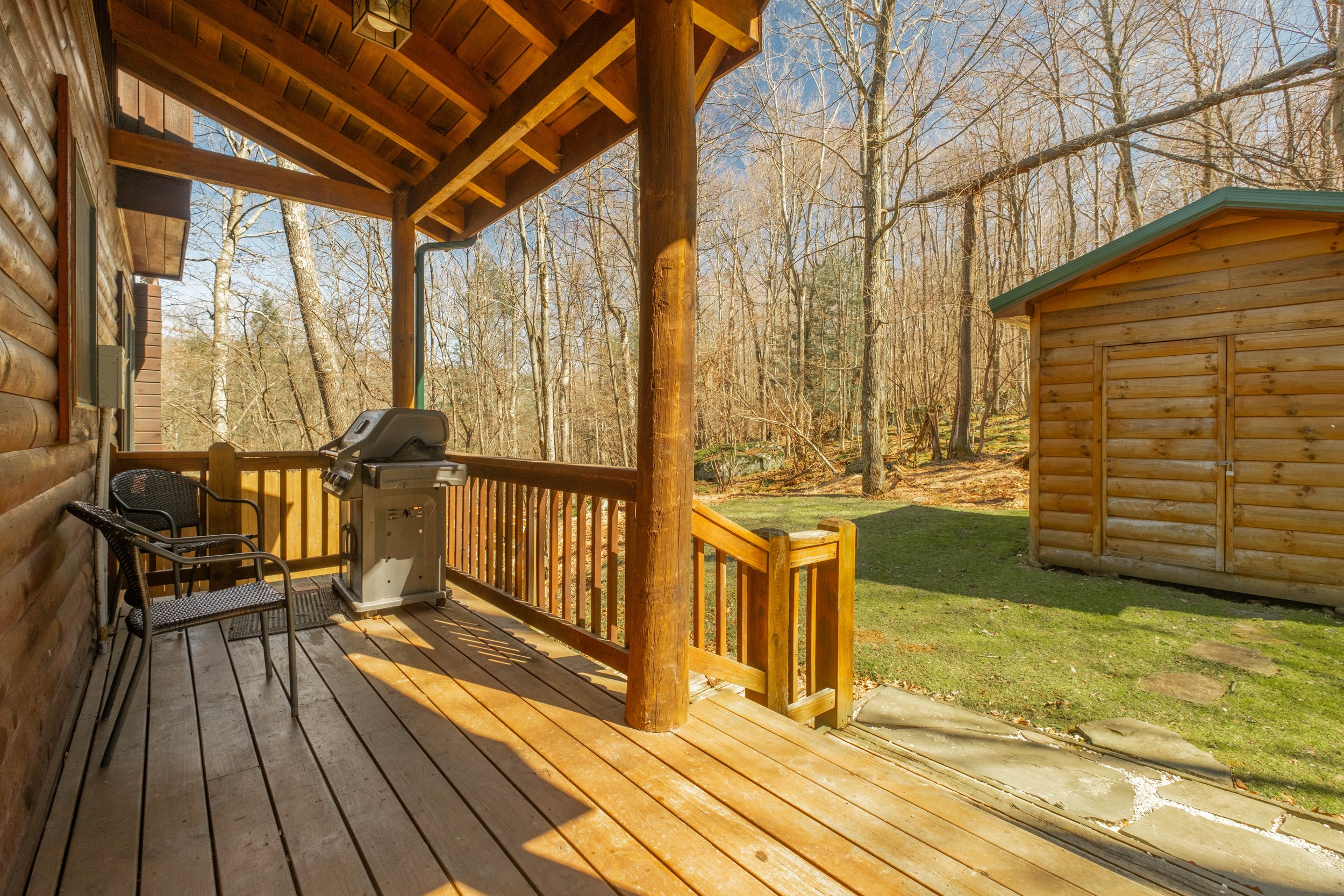 Wooden porch with outdoor grill and chairs, overlooking a grassy yard and wooded trees.