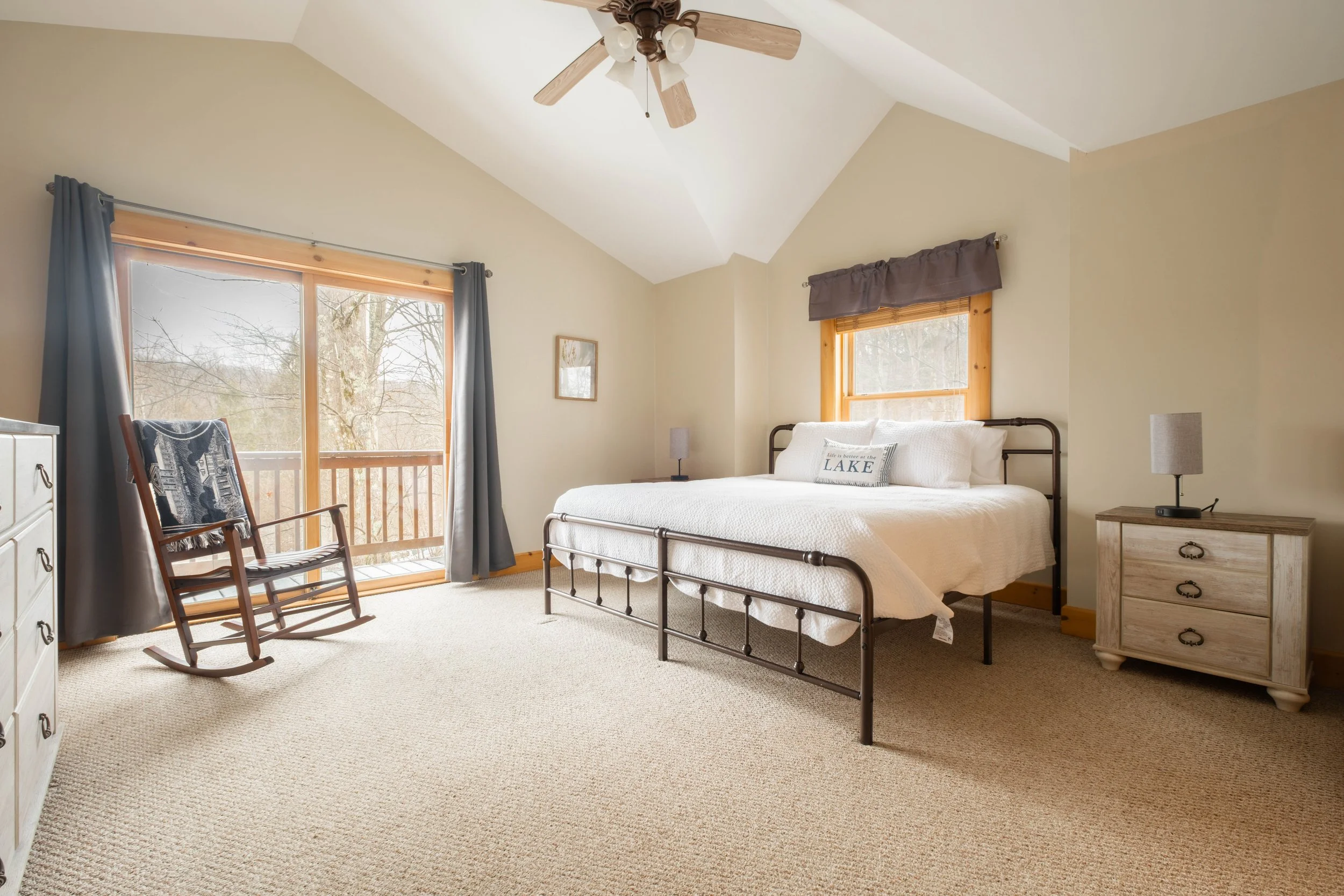 A bedroom with a white bed, wooden accents, a ceiling fan, a rocking chair, a window with curtains, and a sliding glass door leading to a balcony, with trees outside.