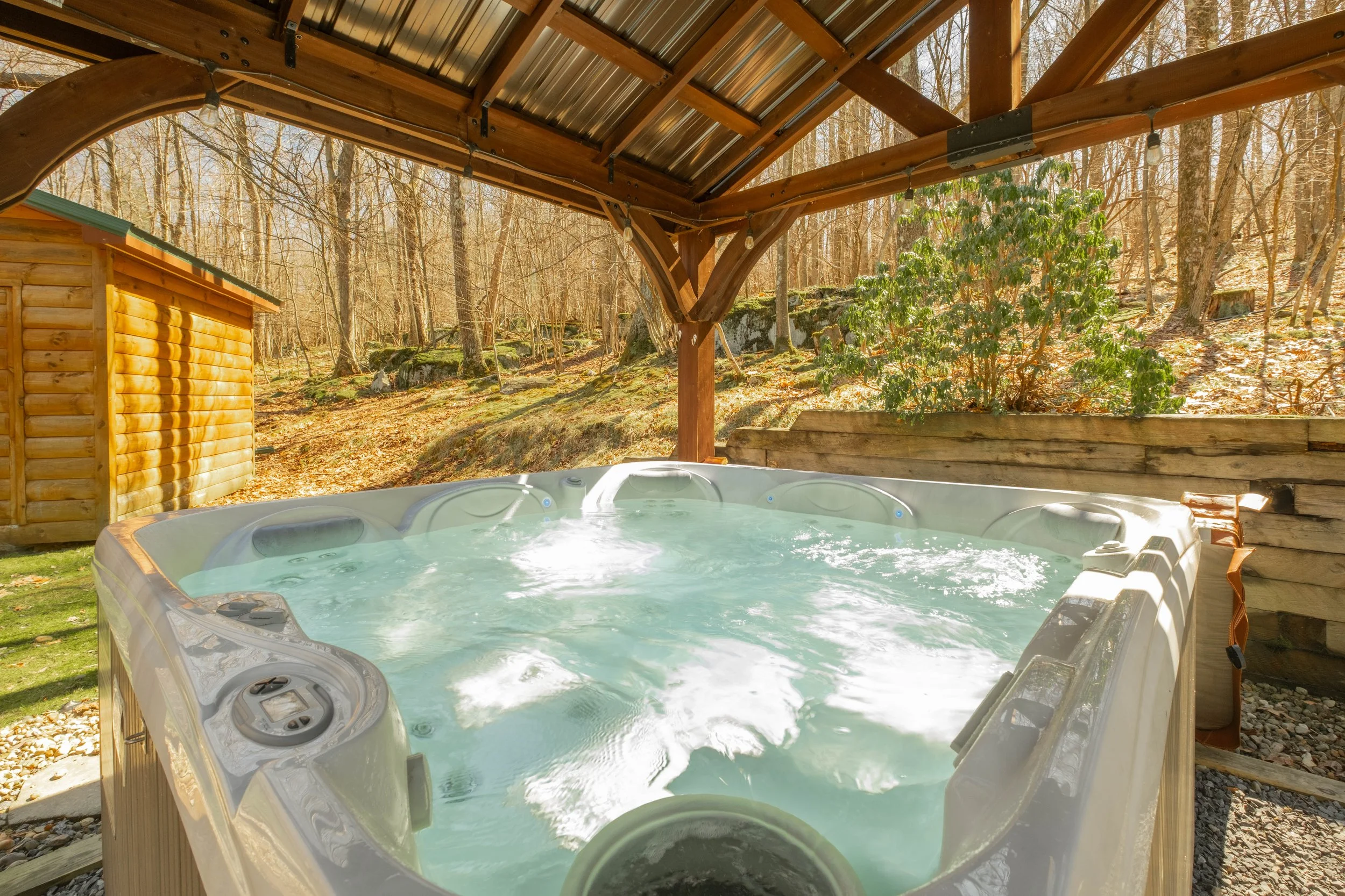 Outdoor hot tub on a wooden deck with a rustic wood shelter, surrounded by trees and rocks in a wooded area during the daytime.